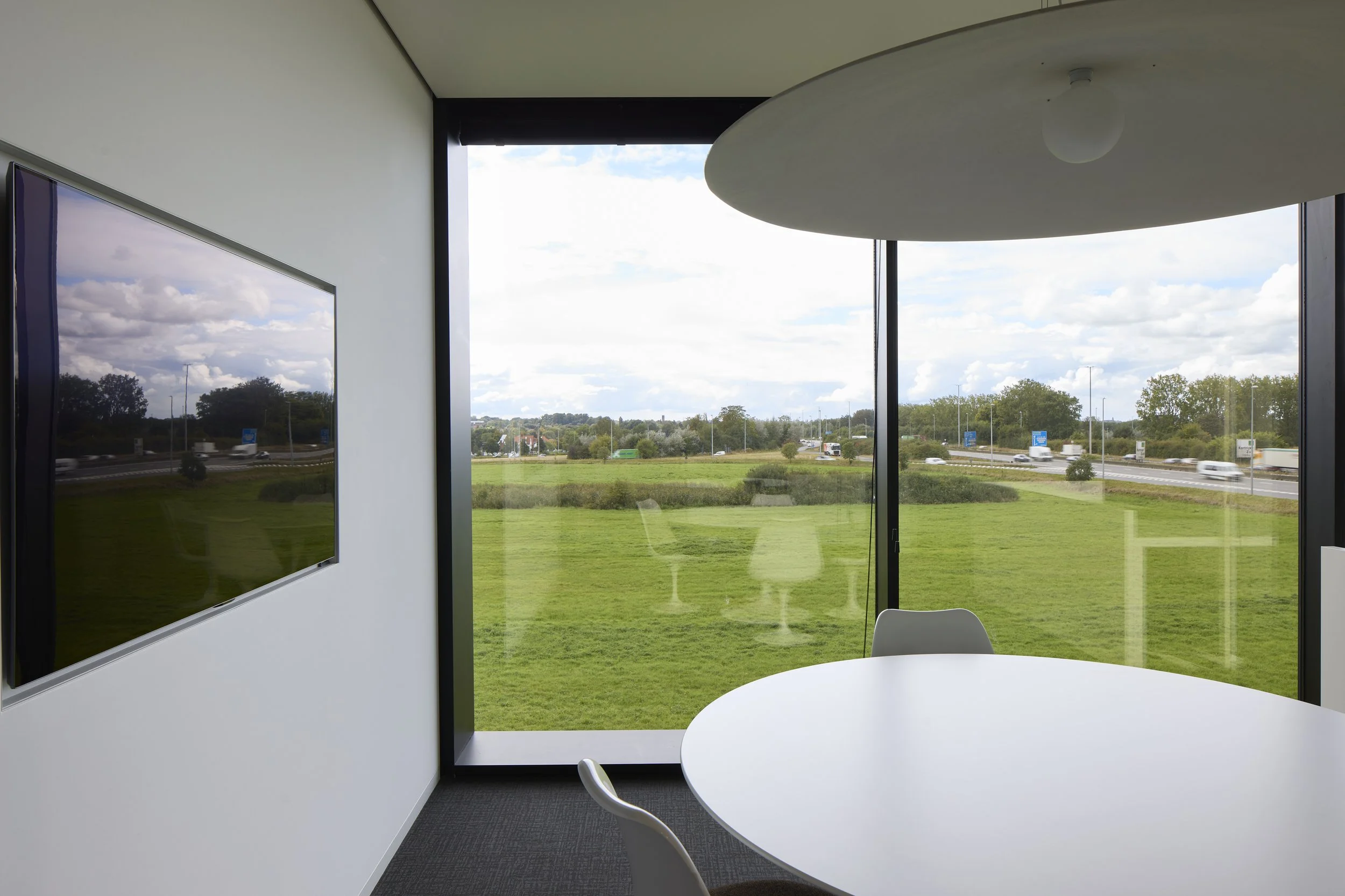 Interior of a modern office with a white round table and chairs, large windows showing a green field and highway outside, and a flat-screen TV mounted on a white wall.