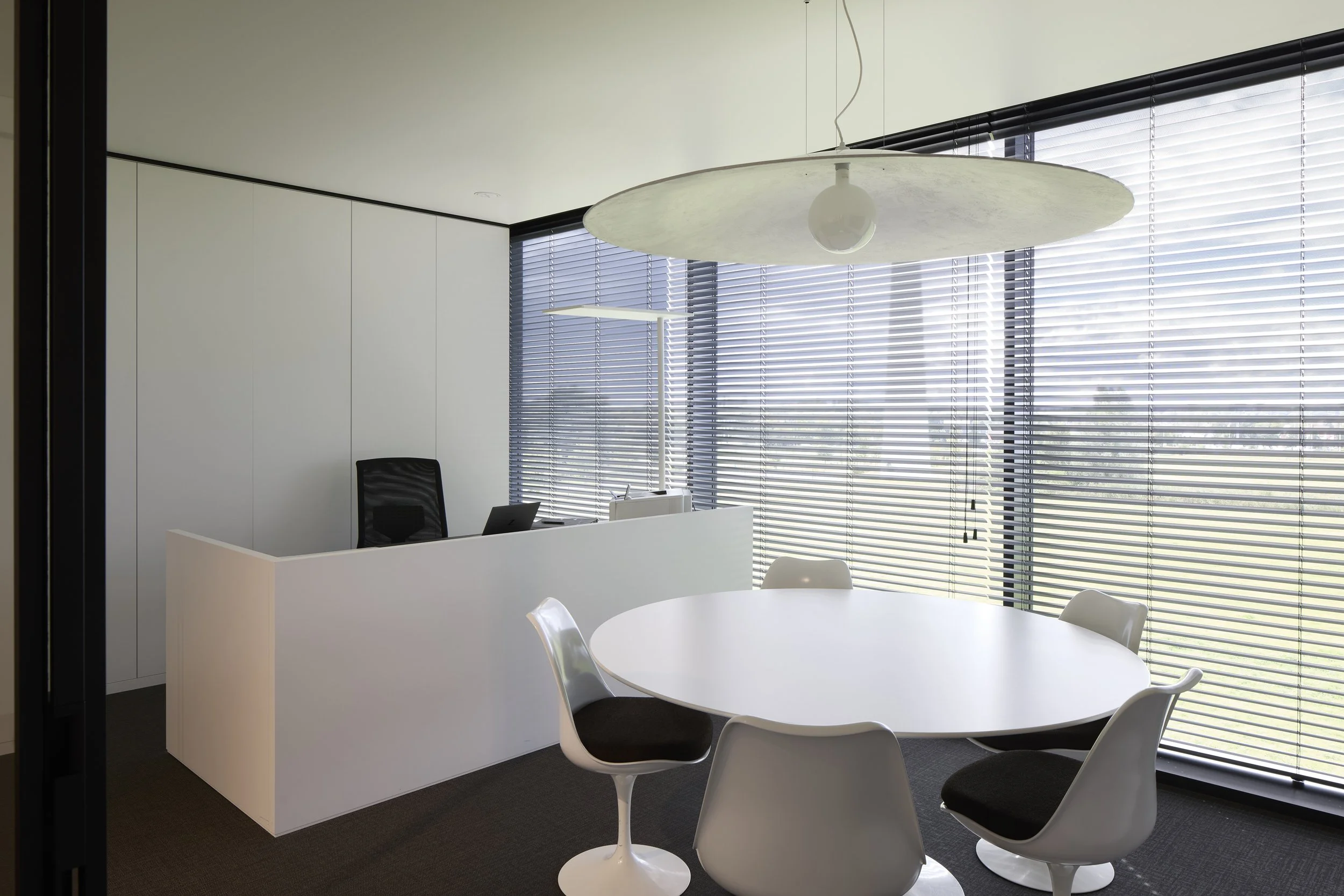 Modern office with white walls, curved white desk, black office chair, white round table, white chairs, large windows with horizontal blinds, and a hanging ceiling light fixture.
