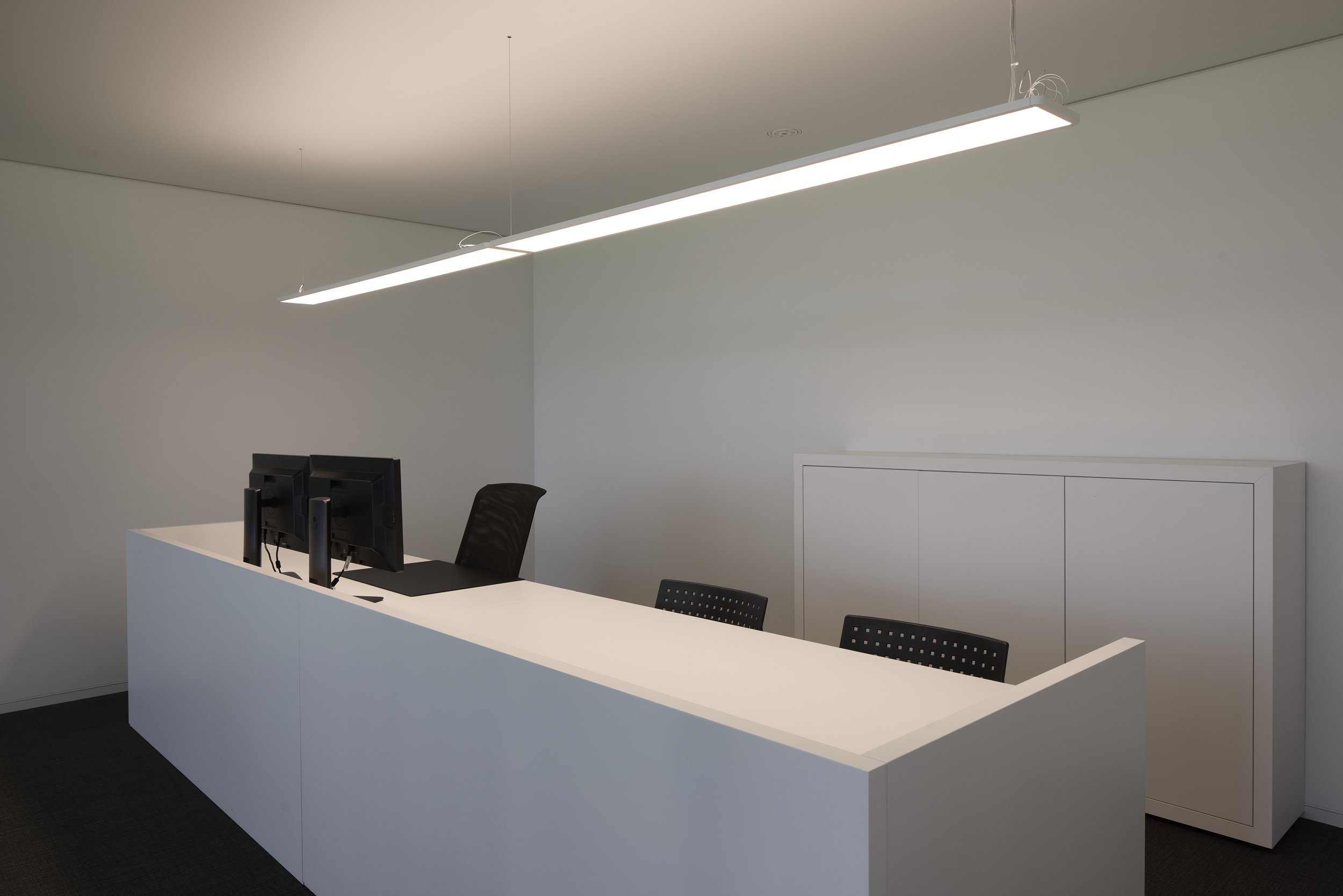 Modern minimalistic office reception desk with two computer monitors and black chairs, white walls, and an elongated ceiling light fixture.