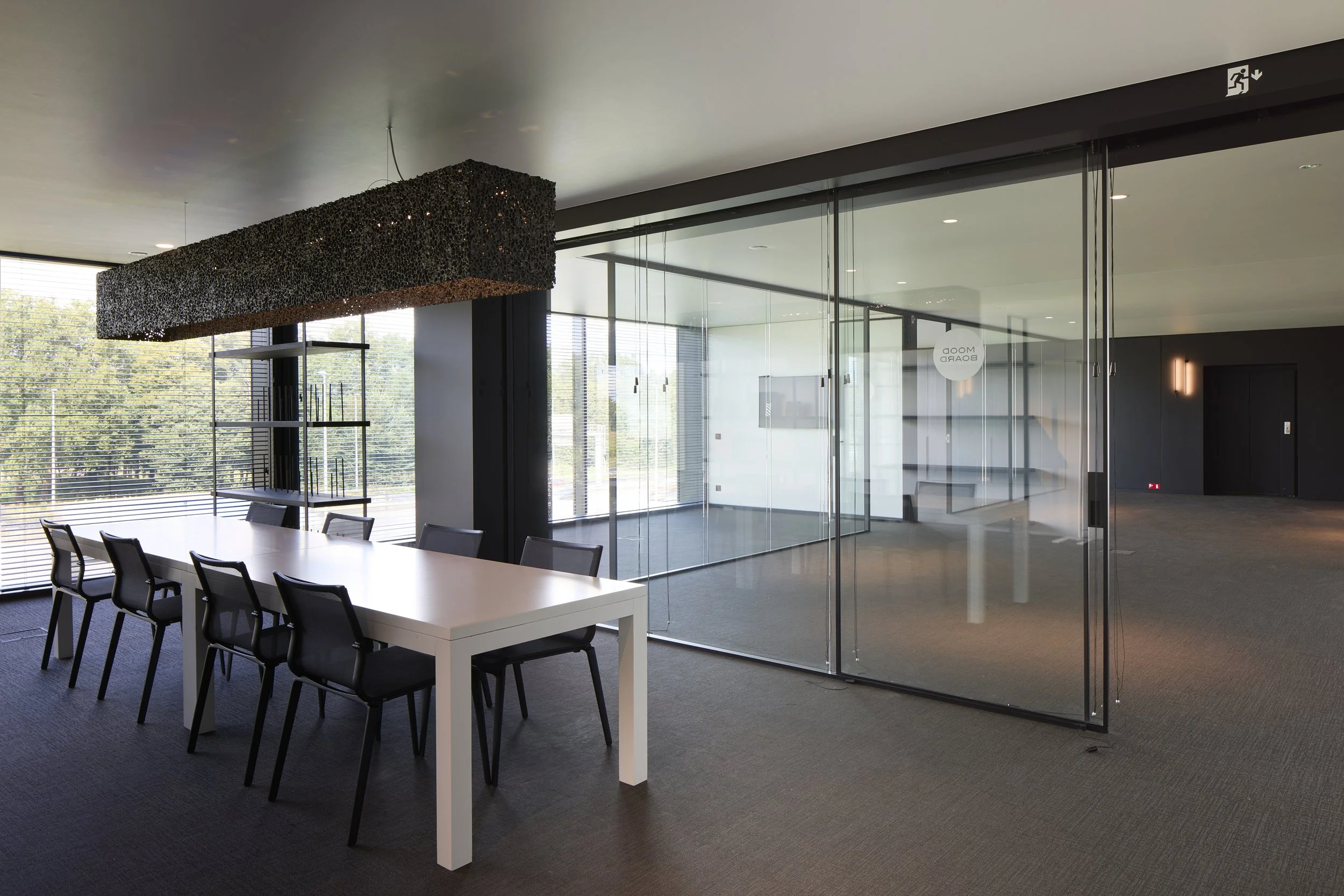 Modern conference room with a long white table, black chairs, large windows with blinds, and a glass partition with the words 'ROOM BOARD' on it, interior lighting, and a view of trees outside.