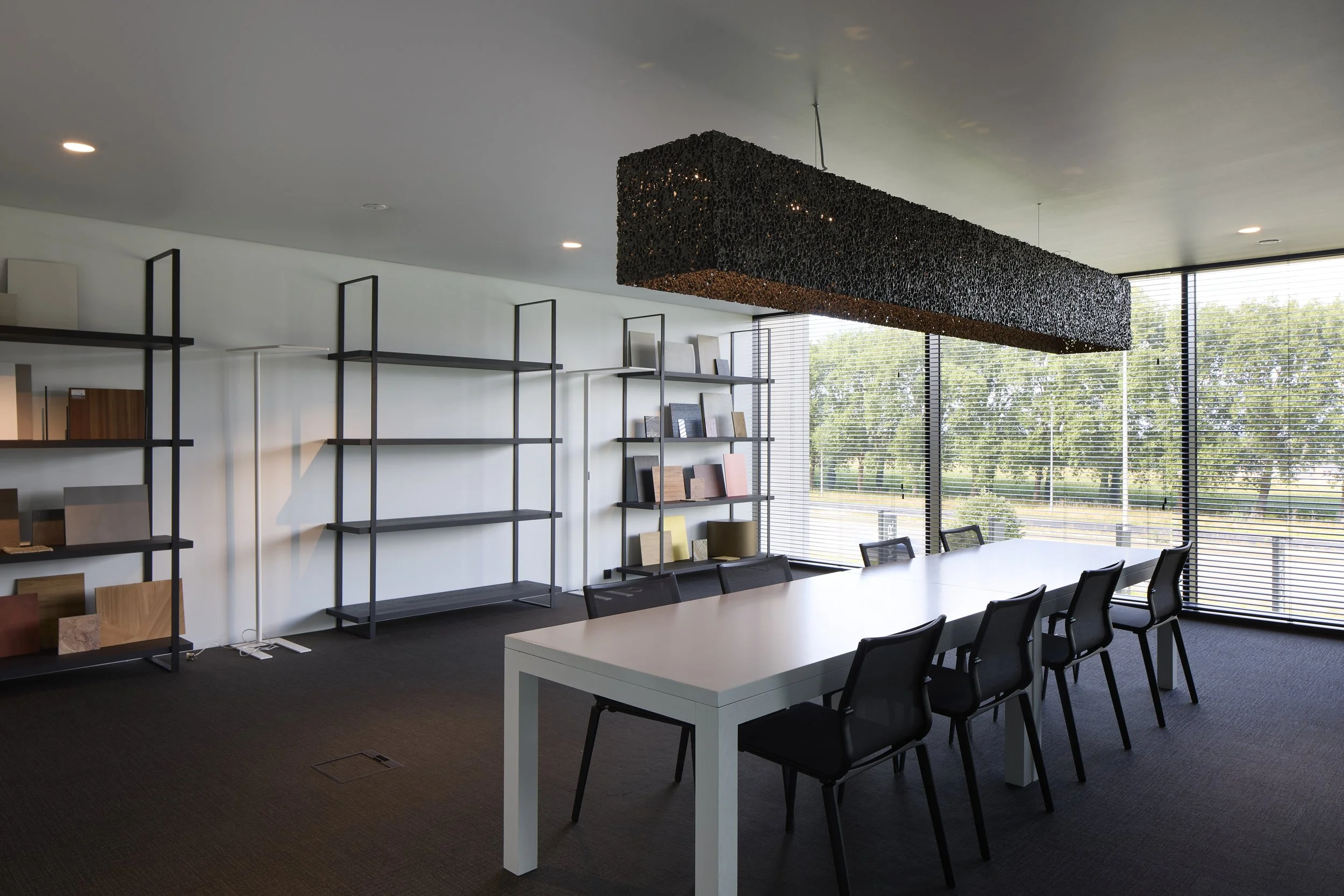 An office conference room with a large white table, black chairs, a black decorative ceiling light, and large windows with blinds overlooking trees outside.
