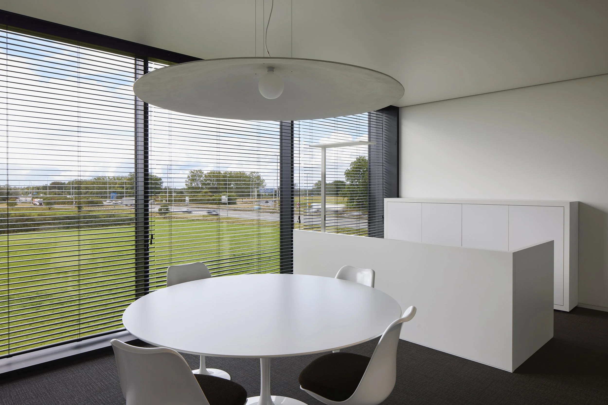 Modern conference room with a round white table and five white chairs, large windows with horizontal blinds showing a parking lot and trees outside, white wall-mounted cabinet, ceiling light, and a bright sky with clouds.