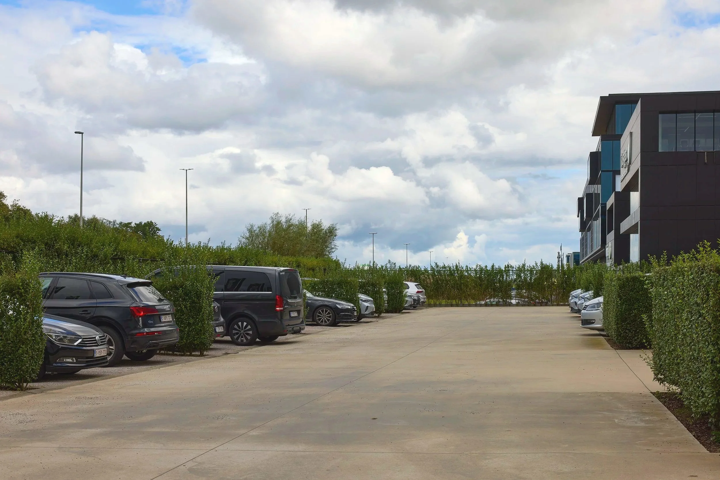 A parking lot with several parked cars and bushes along the sides. In the background, there is a modern building with large windows and a cloudy sky overhead.