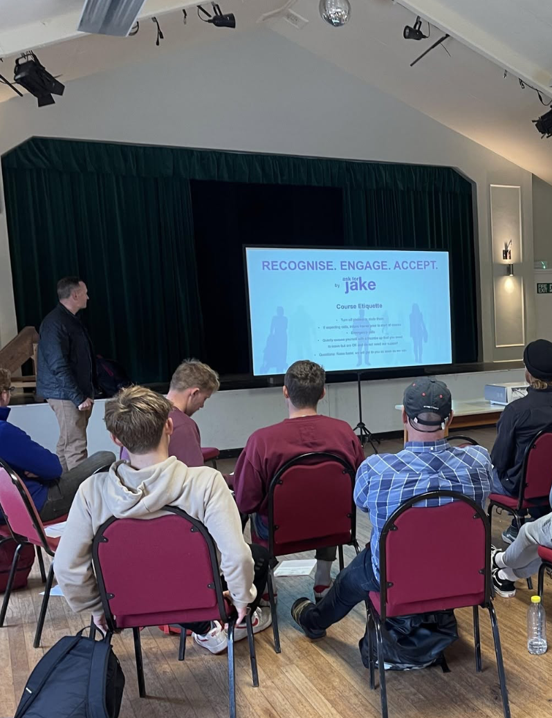 A presentation in a conference room with a screen displaying a slide titled 'Recognise. Engage. Accept.' featuring silhouettes of people and the name 'Jake.' Six people are seated facing the screen, with one person standing to the side.