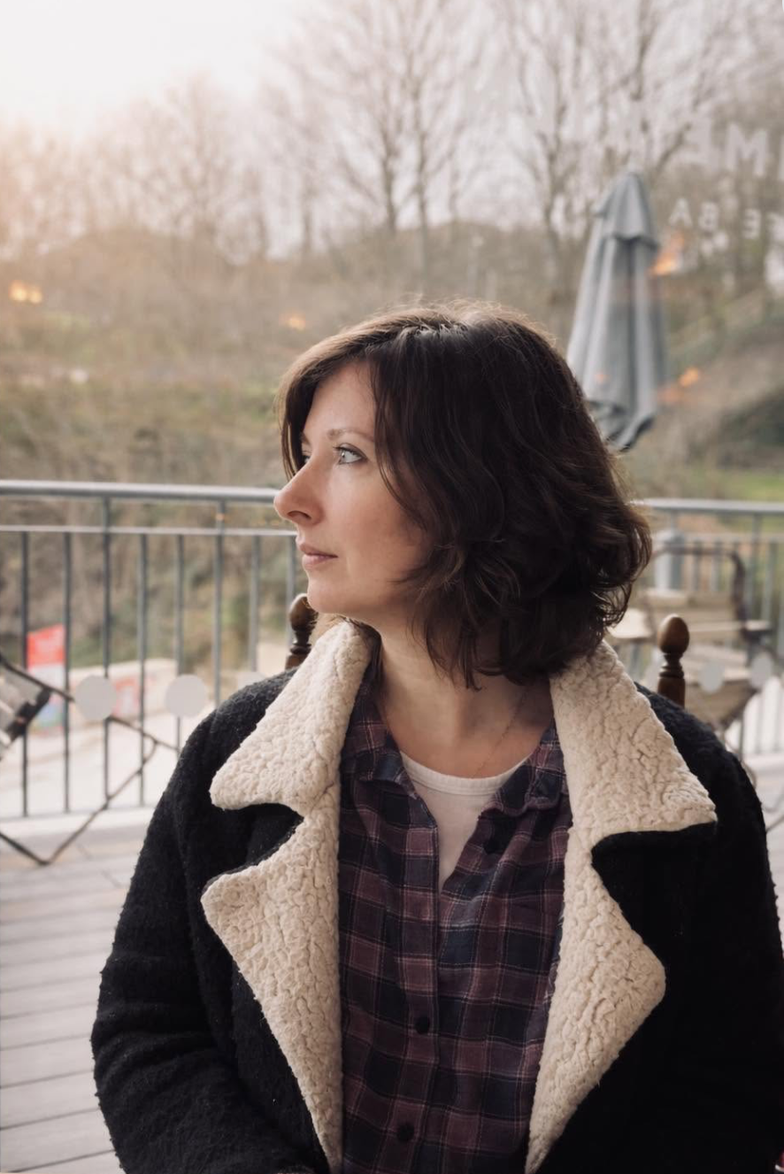 A woman with dark brown, curly hair looking to her left, sitting outside on a wooden deck with a metal railing, trees in the background, and a gray umbrella