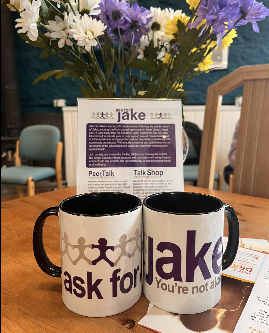 Two white coffee mugs with purple and gray text, one says "ask for Jake" and the other says "you're not alone," placed on a wooden table with a brochure and a flower arrangement in the background.