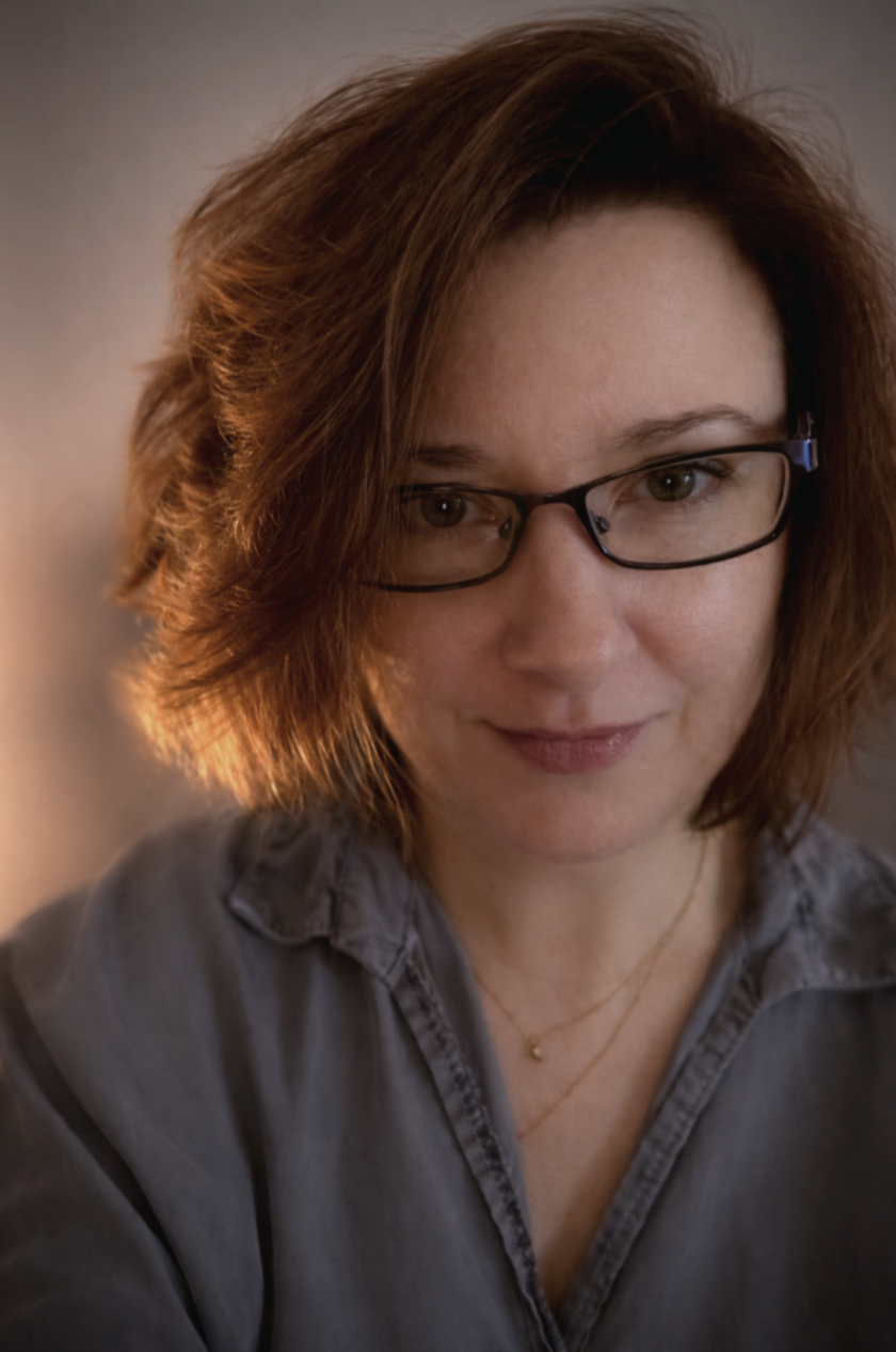 A woman with red hair, glasses, and a gray shirt smiling softly, with soft lighting highlighting her face.