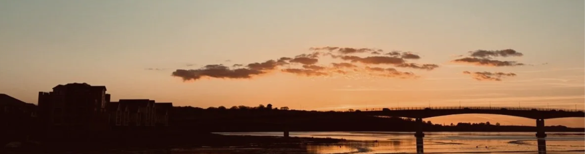 Sunset sky over a river with a bridge and silhouette buildings on the left.