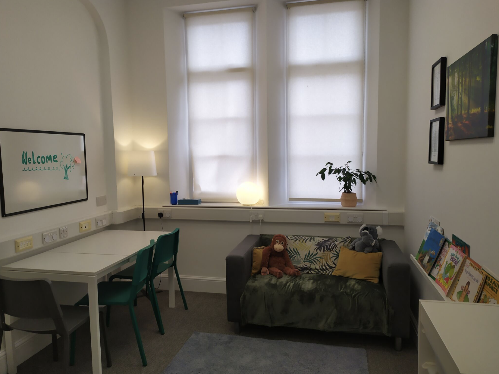 A room with two large windows with white blinds, a white table with green chairs, a sofa with stuffed animals, a plant, and a bookshelf with children's books.