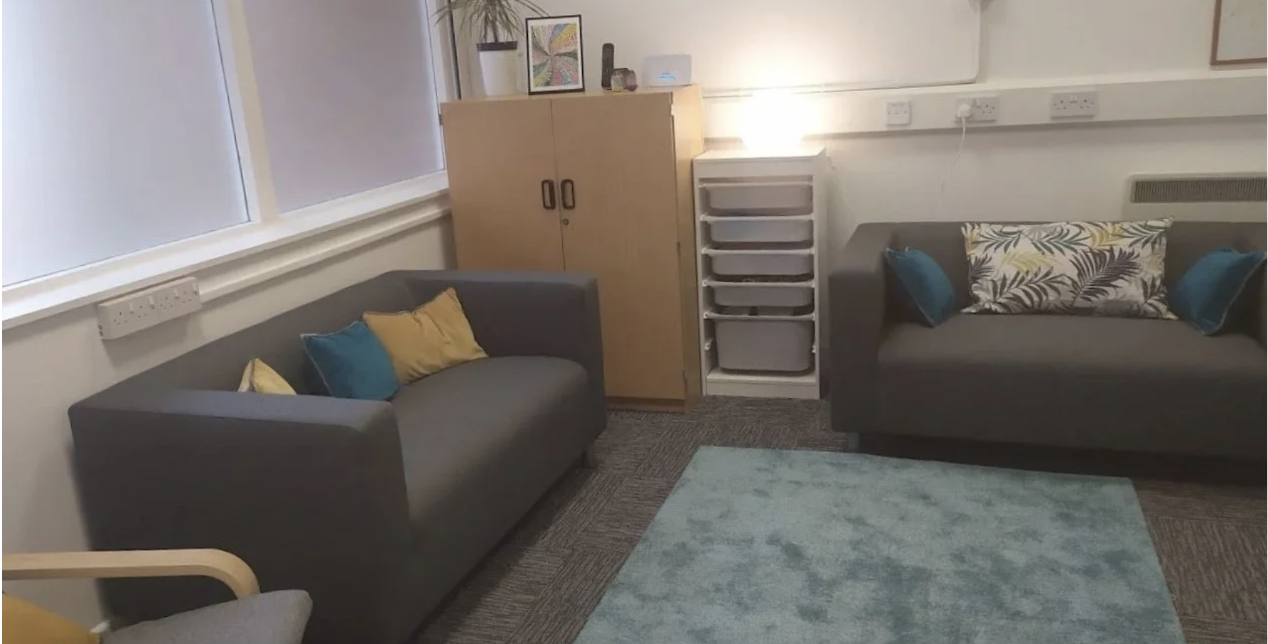 A living room with two gray sofas, each adorned with colorful cushions in yellow, blue, and patterned designs. A wooden cabinet and a white plastic drawer unit are situated between the sofas and a wall with electrical outlets. There is a window with closed blinds on one side and a light blue rug on the carpeted floor. Decorations include a framed colorful abstract artwork, potted plant, and small items on the cabinet.