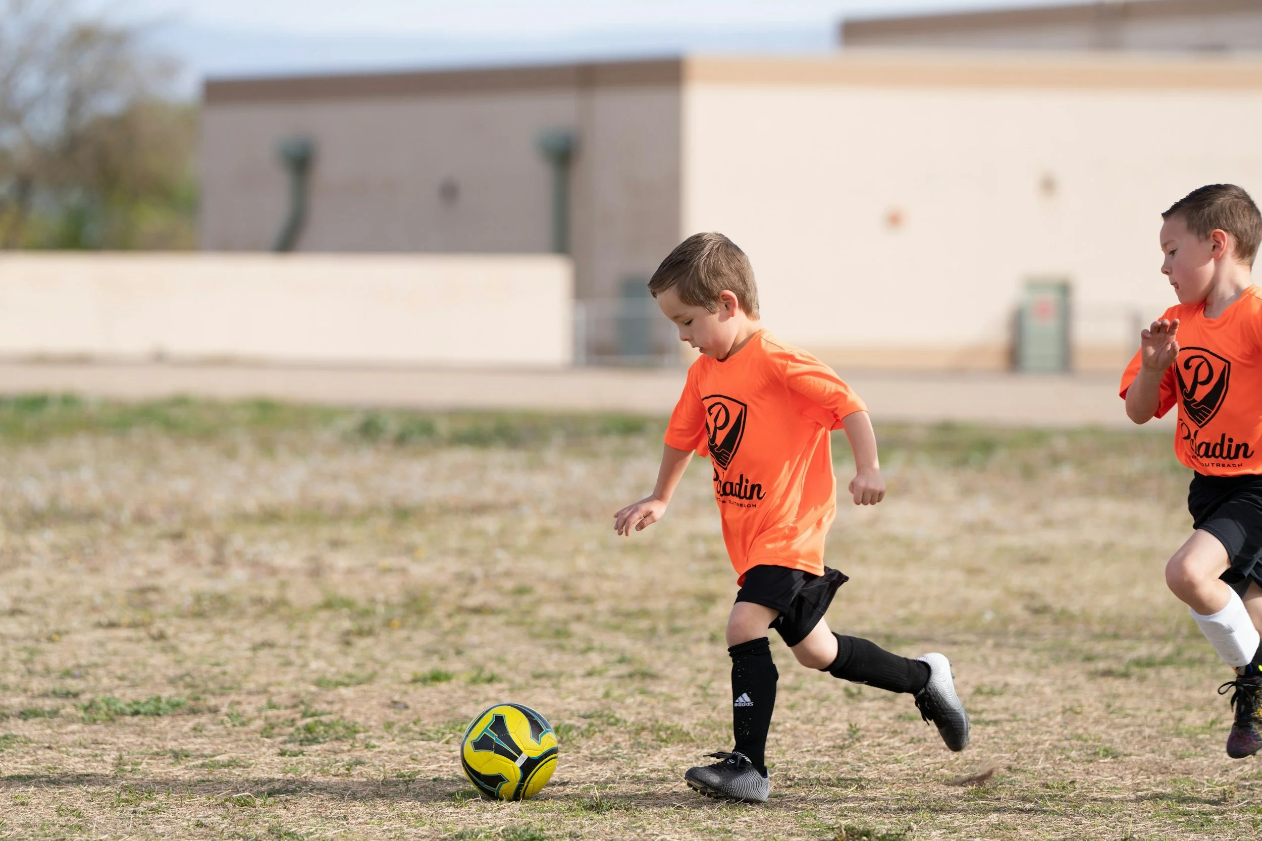 Two young boys playing soccer outdoors, wearing orange shirts and black shorts, chasing a yellow and black soccer ball on a grassy field with a building in the background.