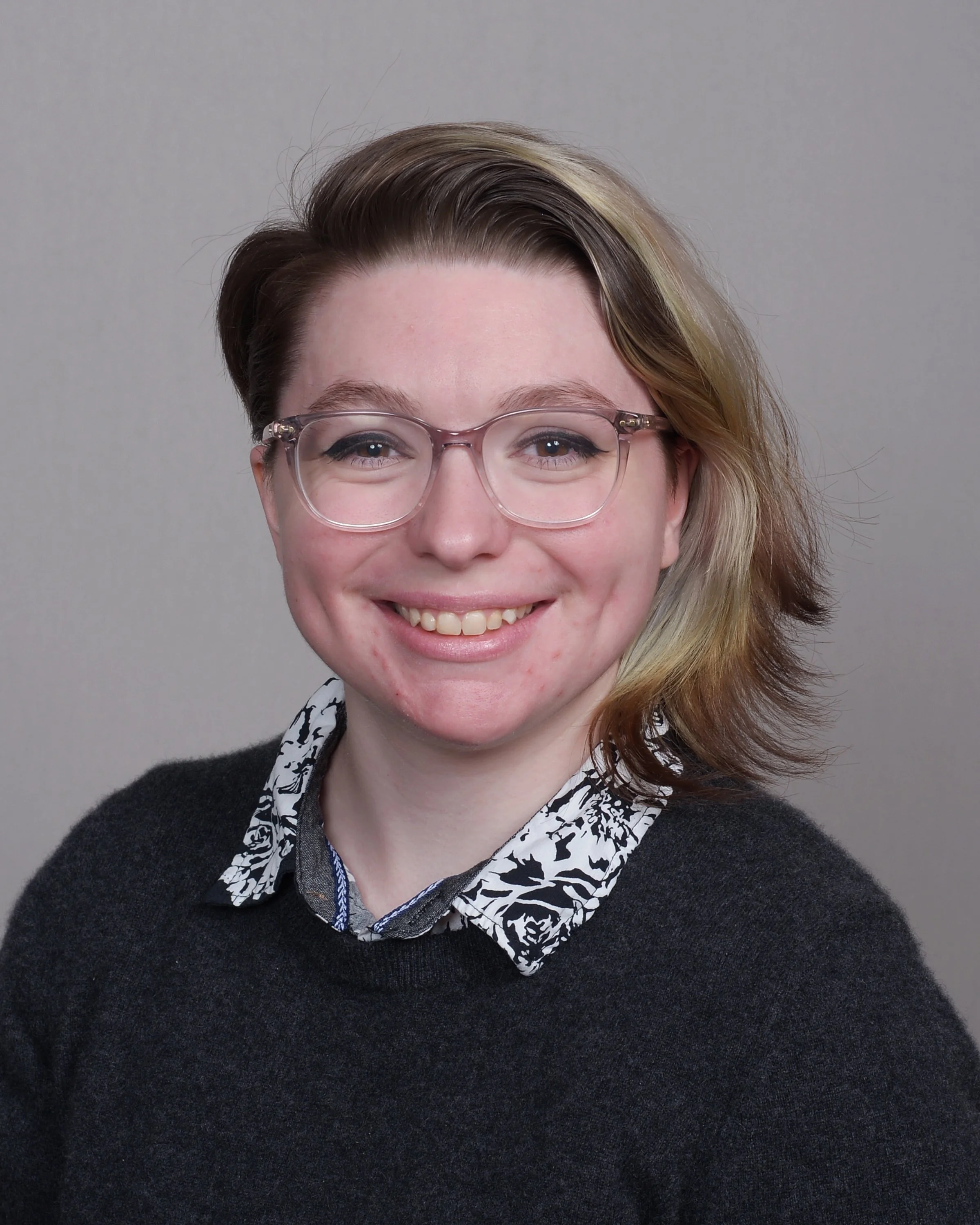 Close-up of a smiling woman with light skin, short hair with dark and blonde highlights, wearing glasses, a black sweater, and a collared shirt with a black and white pattern, against a plain gray background.