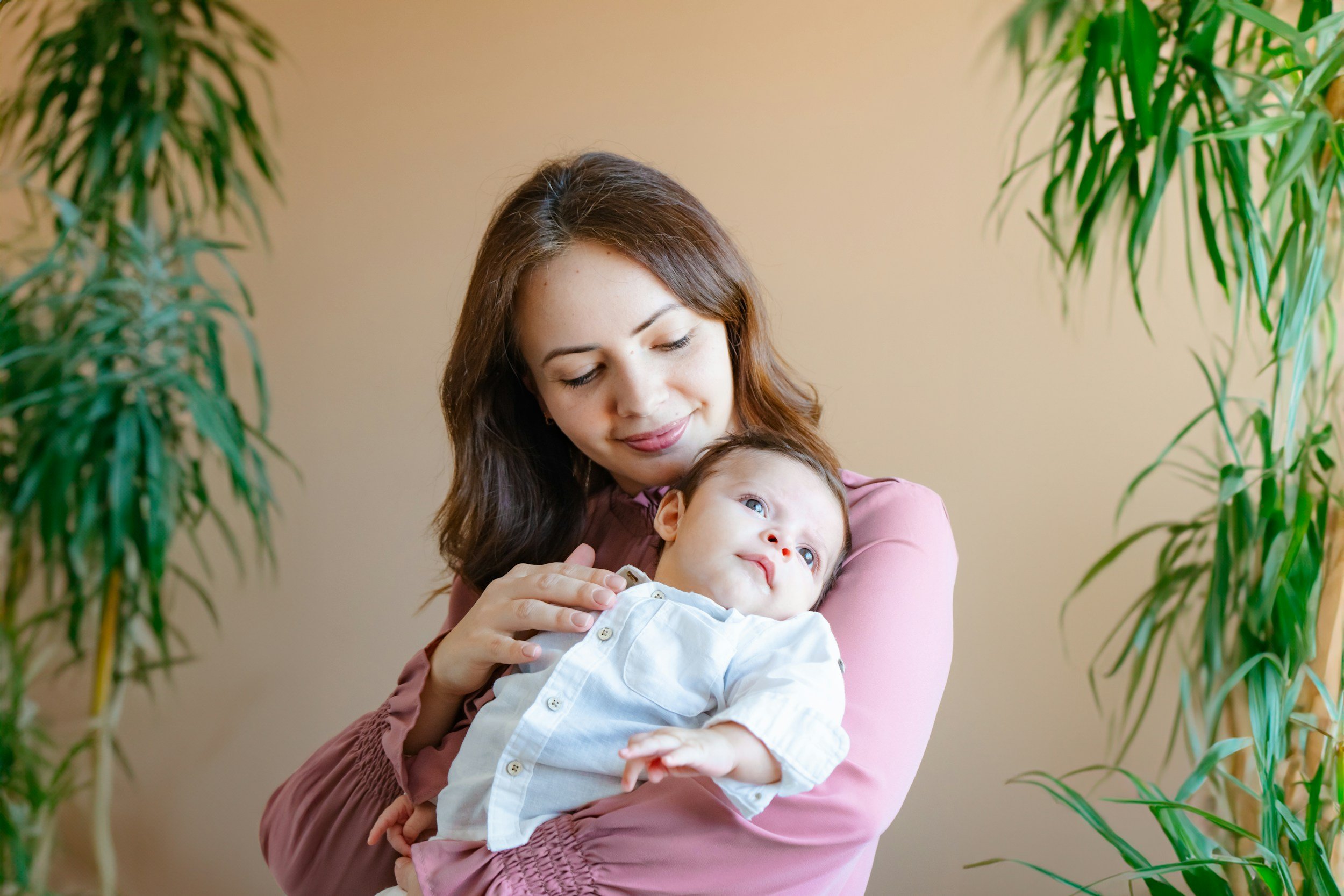 A woman holding a young child in front of a beige wall, with green plants on each side. The woman has long brown hair and is wearing a mauve top. The child has blonde hair, blue eyes, and is dressed in a white shirt.