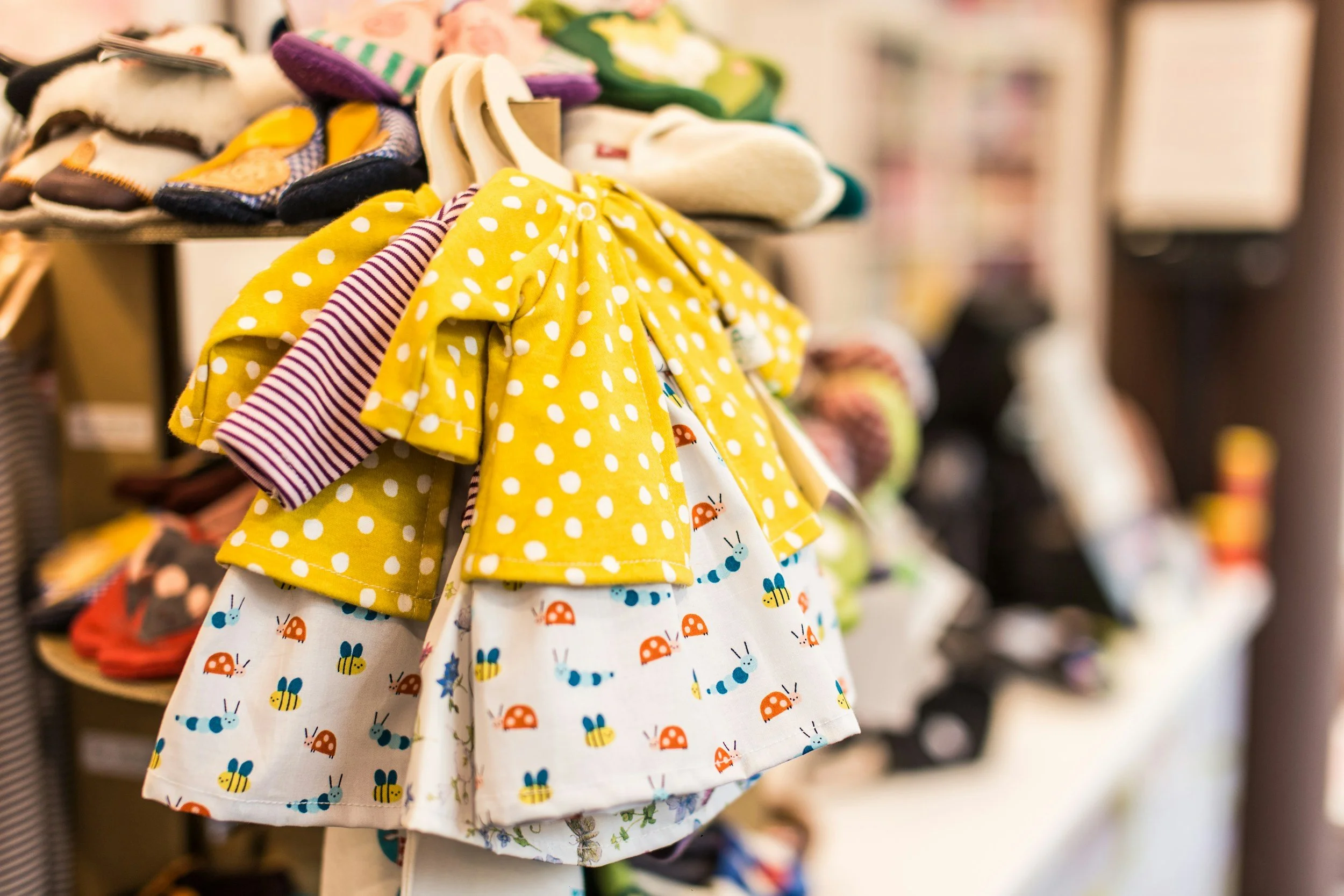 A display of colorful children's clothing, including a white dress with ladybug and bumblebee patterns and a yellow dress with white polka dots, hanging on a rack in a store.
