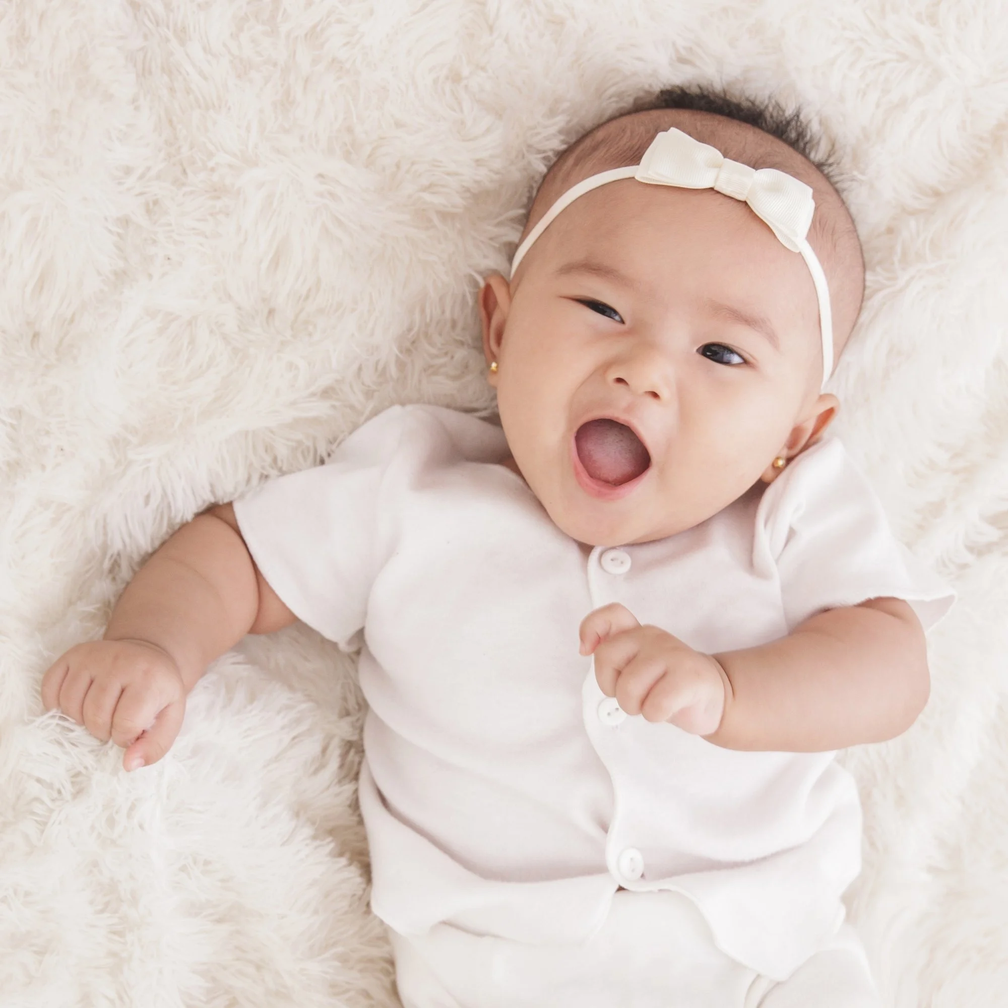 A smiling baby lying on a soft, fluffy white blanket, wearing a white short-sleeve shirt with buttons and a cream headband with a bow.