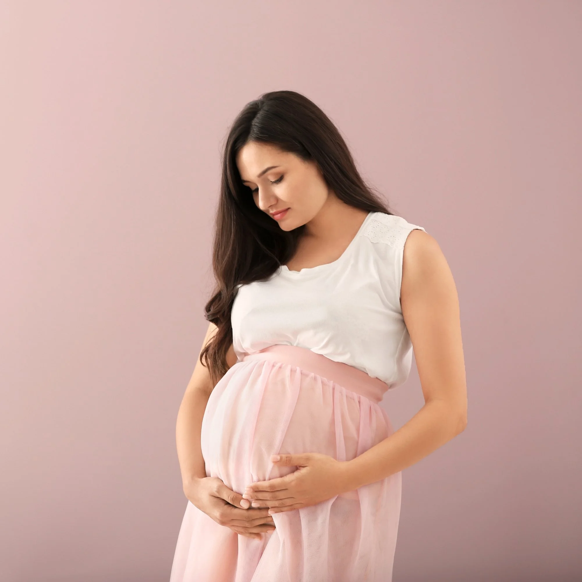 Pregnant woman wearing a white top and pink skirt, gently holding her belly, standing against a pink background.