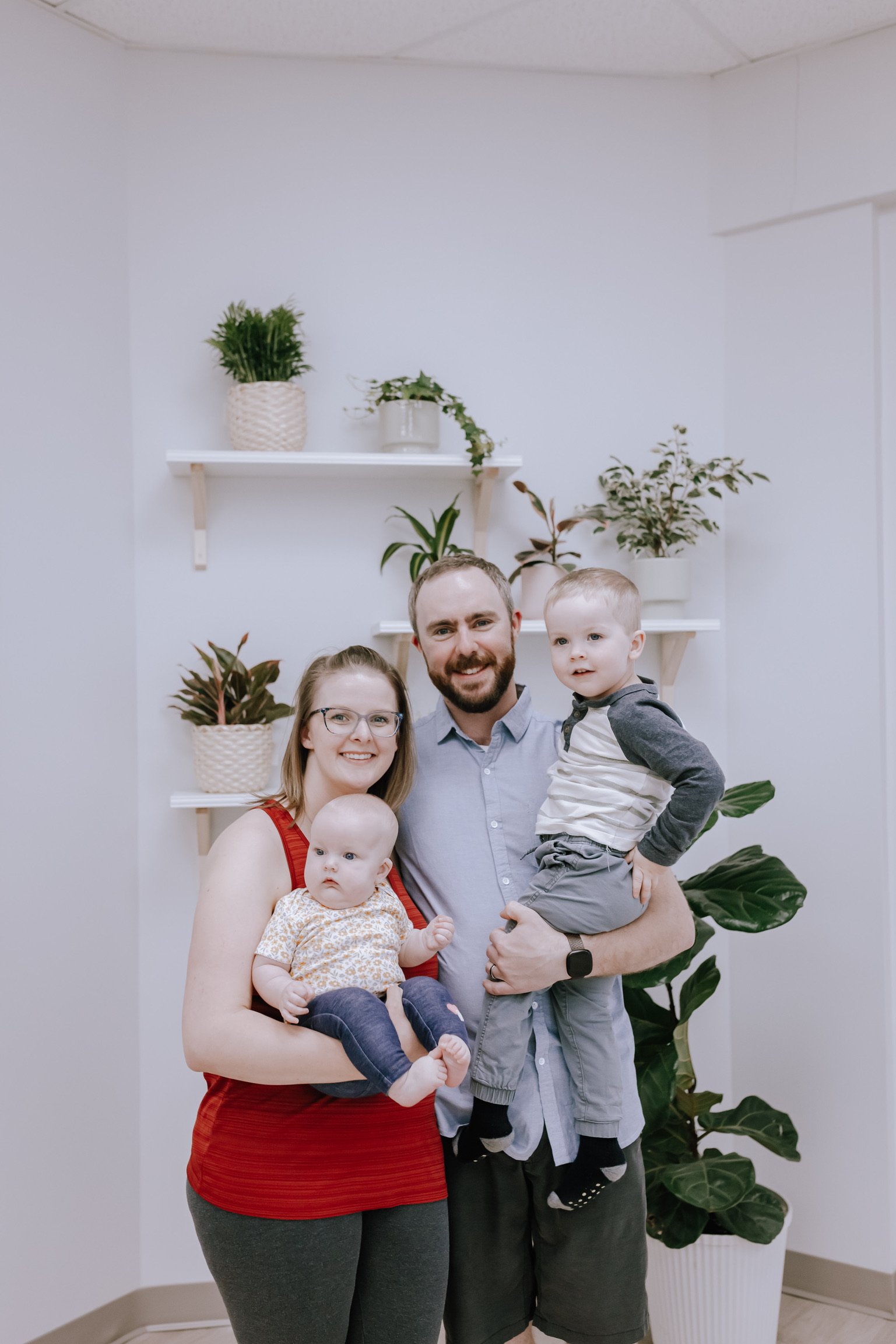 Family of four, overcast lighting, standing in front of white wall with three white shelves holding houseplants, including a large leafy plant and smaller potted plants, with a woman wearing glasses, a red sleeveless top, a man with a beard in a light blue shirt, and two children, one a baby in a floral dress and the other a young boy in a gray hoodie, all smiling at the camera.