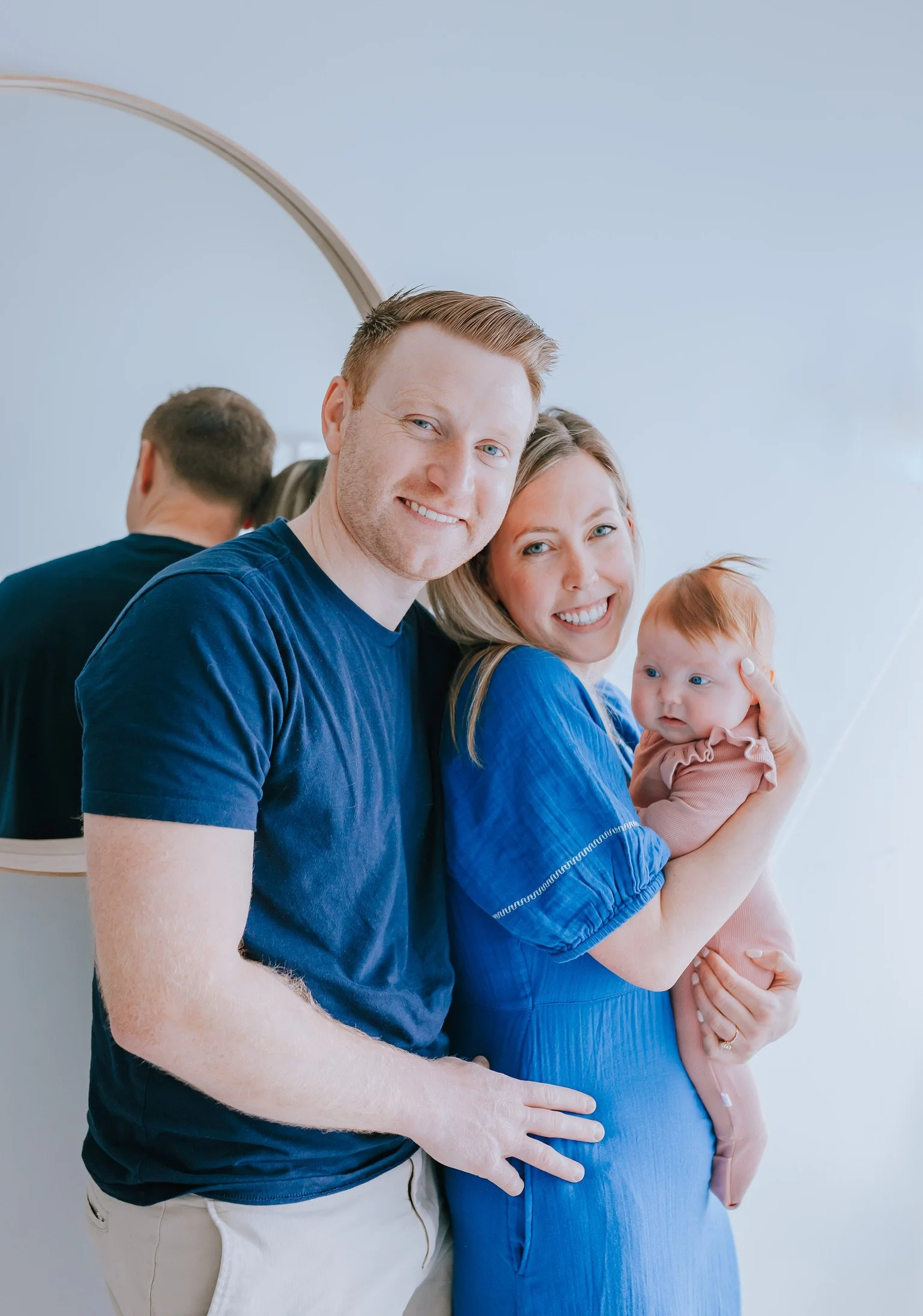 A happy couple, the woman pregnant, with a man standing behind her, and a small child holding her belly, all smiling and looking at the camera.