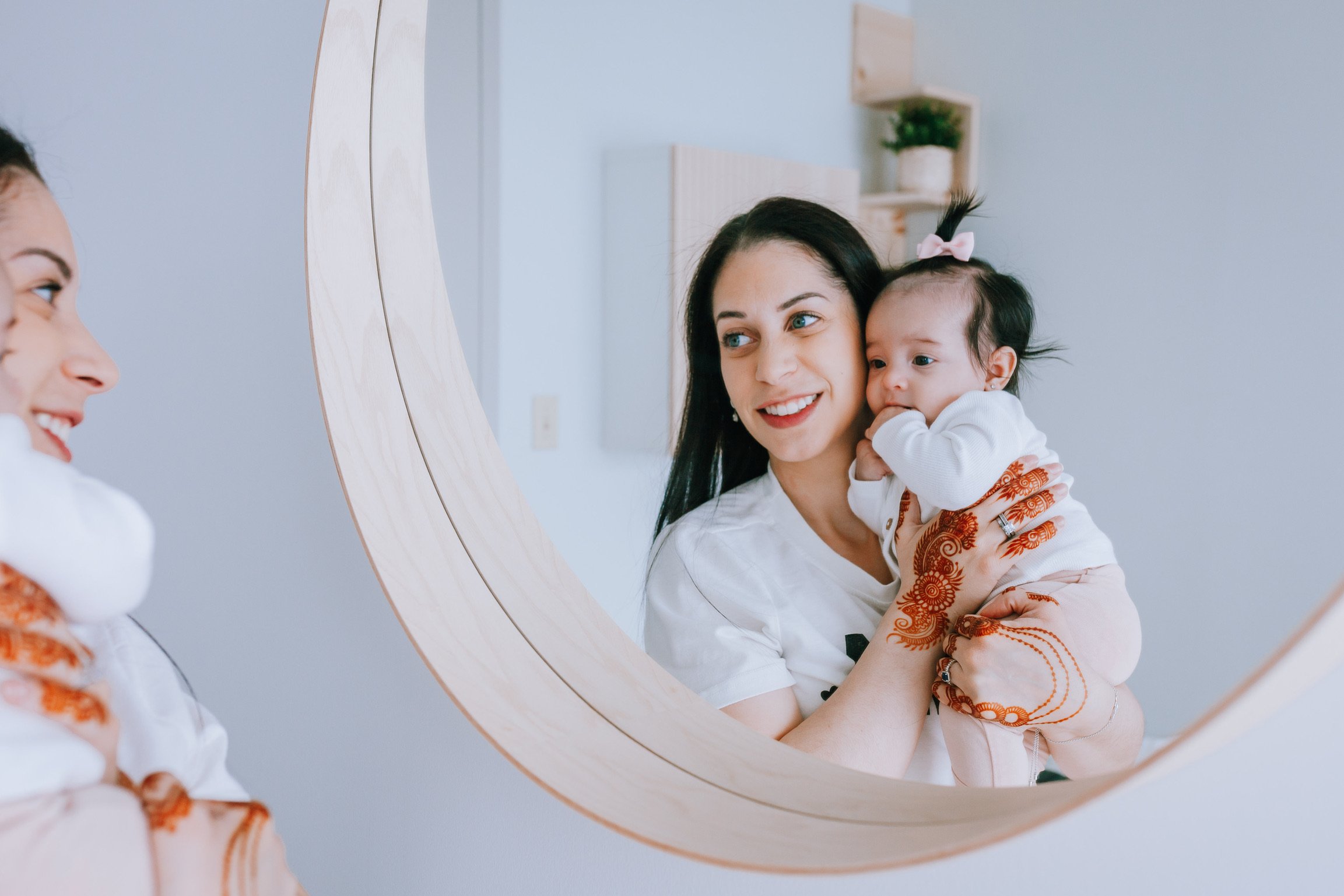 A woman holding a baby girl looking at her reflection in a round mirror, with another woman smiling in the background, in a bright room with light-colored walls and a small wall-mounted shelf with a potted plant.