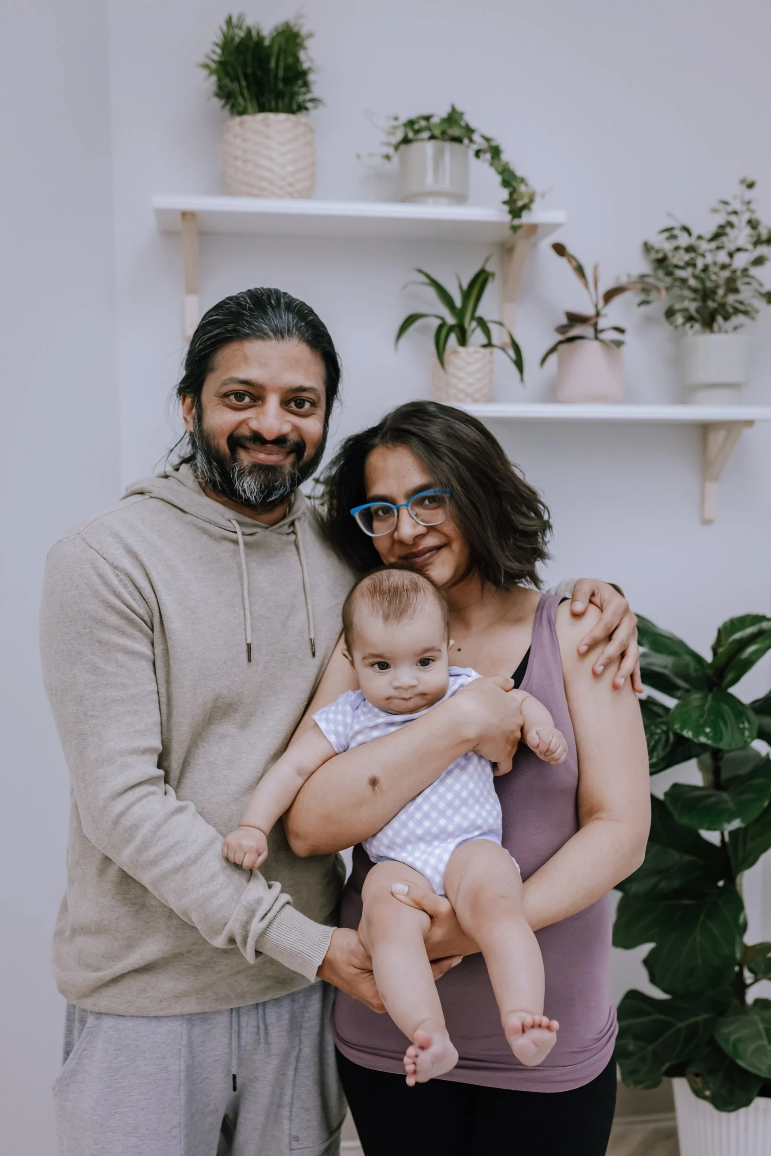 A happy family standing together indoors, holding a baby boy with short hair and wearing a checkered outfit. The man has long dark hair and a beard, wearing a beige hoodie and grey sweatpants. The woman has short dark hair, glasses, and is dressed in a purple sleeveless top, cradling the baby.