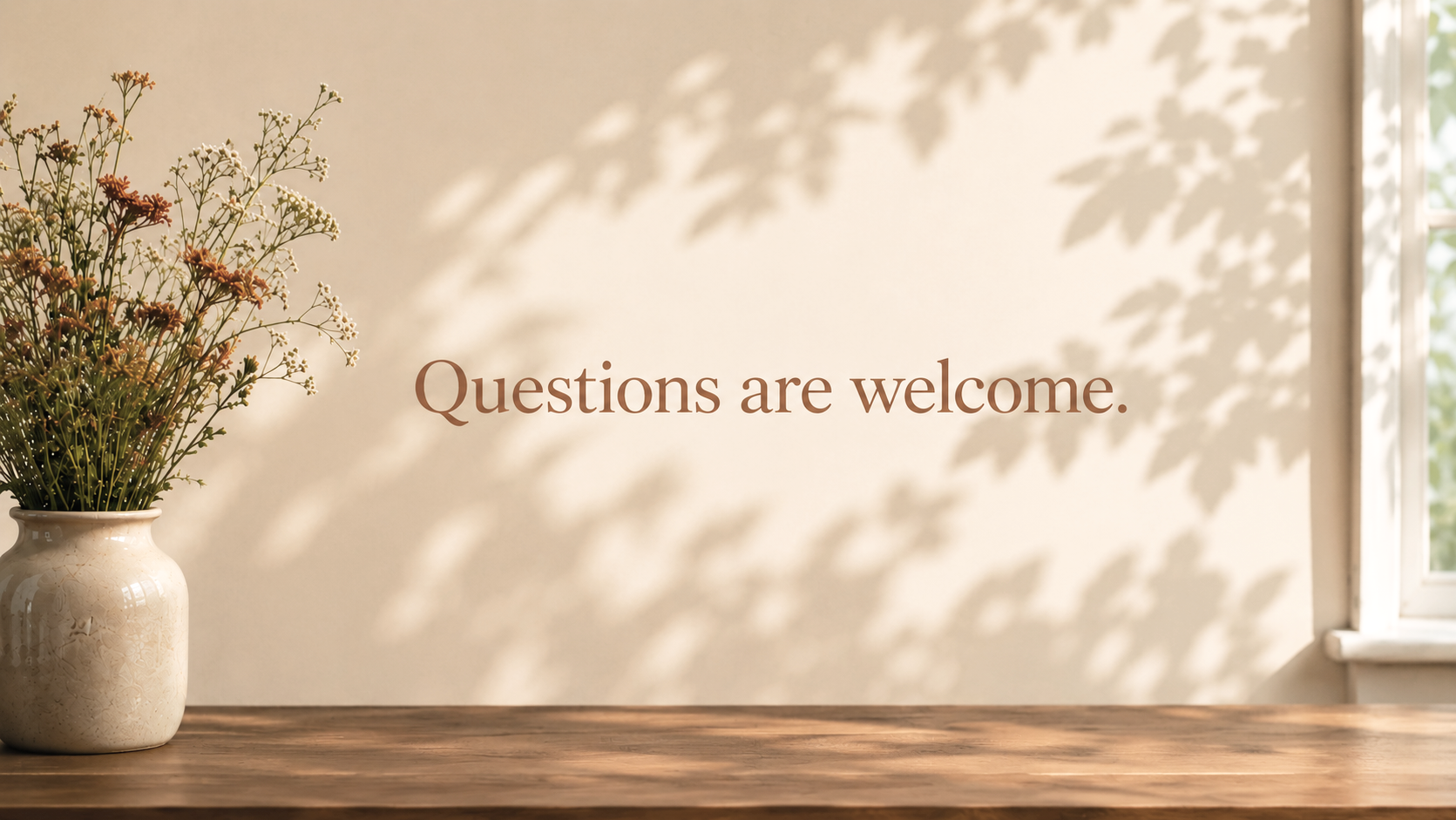 A vase with white and brown flowers on a wooden table, with sunlight casting shadows of leaves on the wall. Text on the wall reads 'Questions are welcome.'