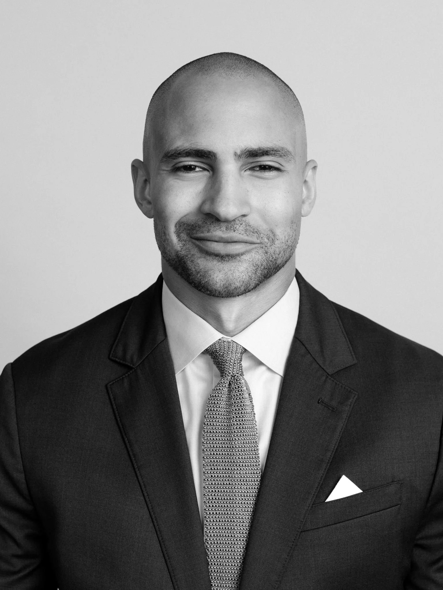 Black and white portrait of a man with a shaved head, wearing a suit, white shirt, and patterned tie, smiling slightly at the camera.