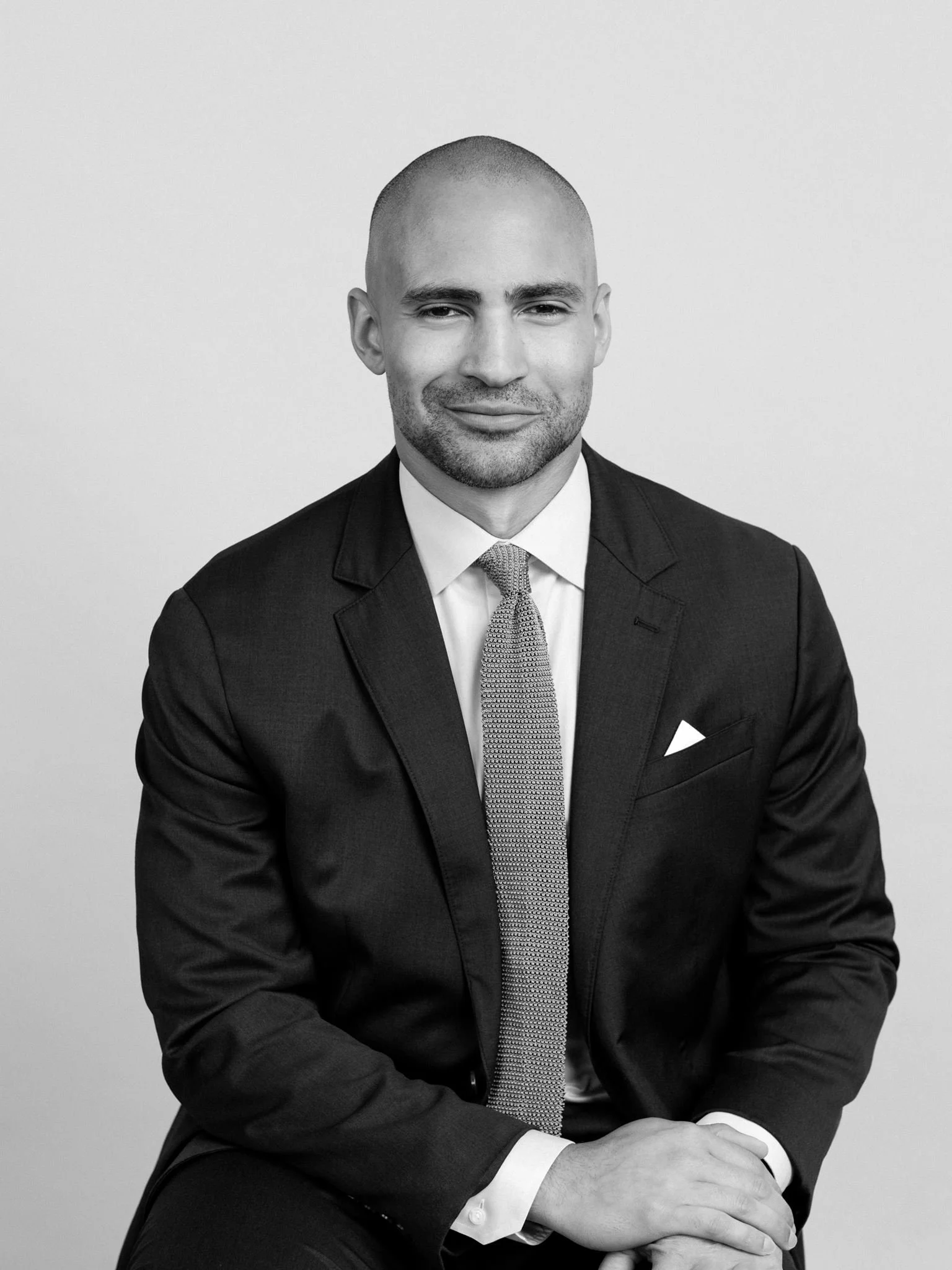 Black and white portrait of a man in a suit and tie, seated with hands folded on his lap, smiling lightly, against a plain background.