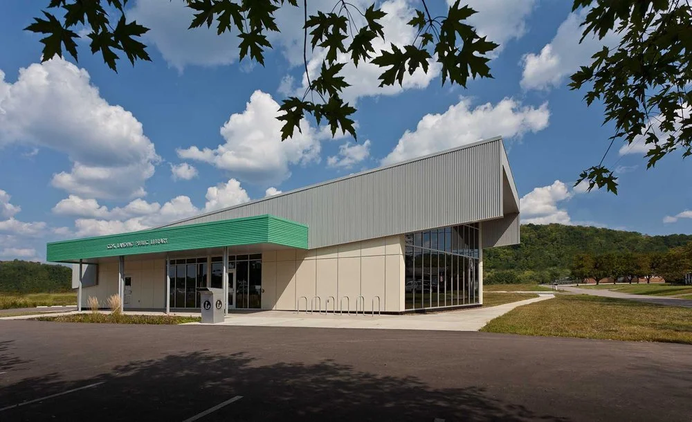 Modern building with green awning, large glass windows, surrounded by a parking lot and grassy area, under a partly cloudy sky.