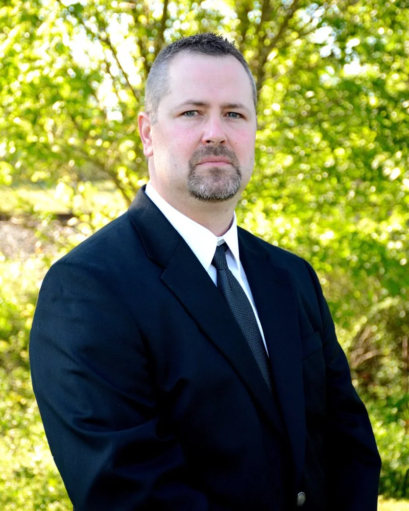 A man in a black suit and tie standing outdoors with green foliage in the background.
