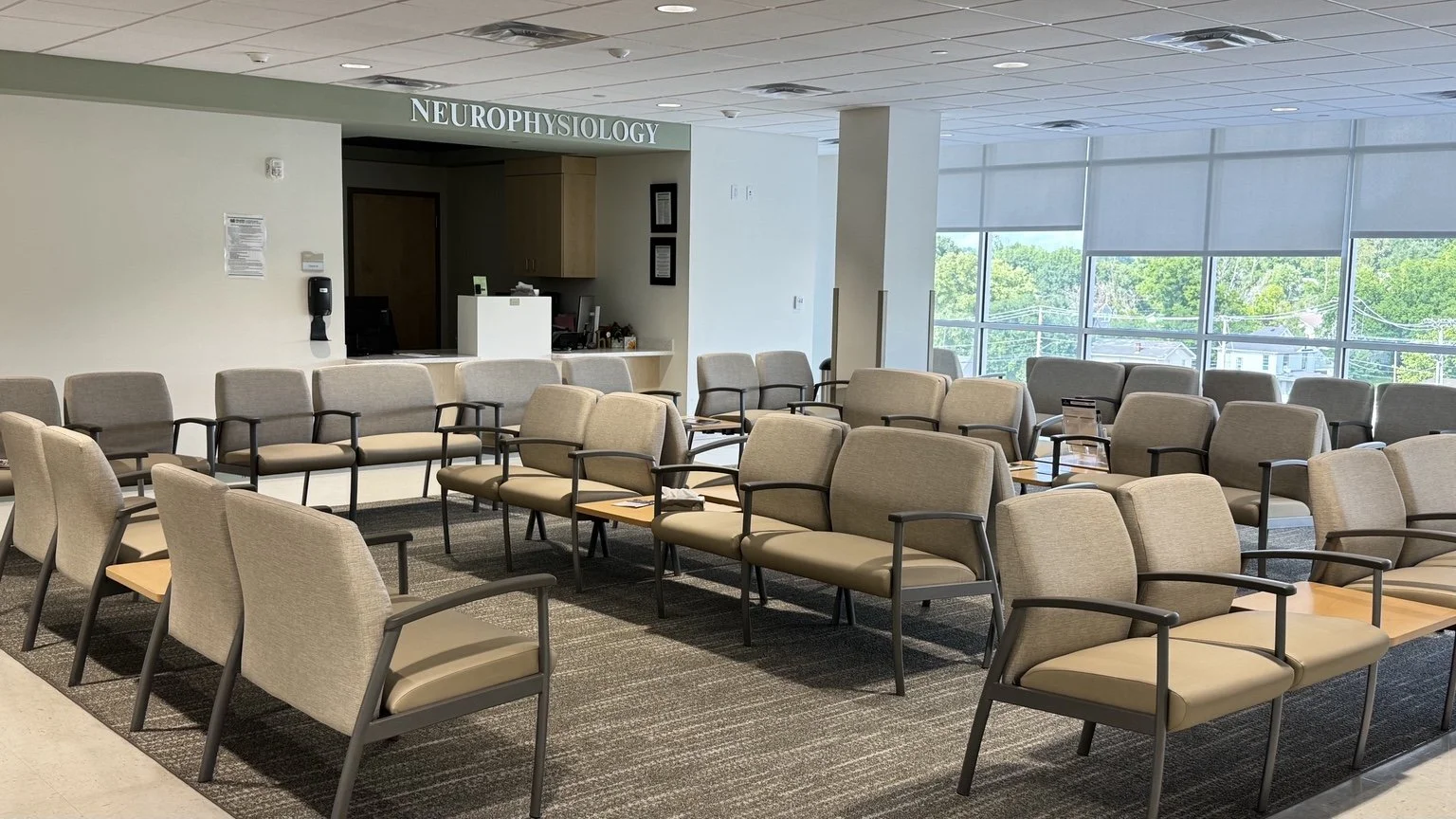Waiting area with rows of beige chairs, large windows with shades, and a sign for neurophysiology in the background.