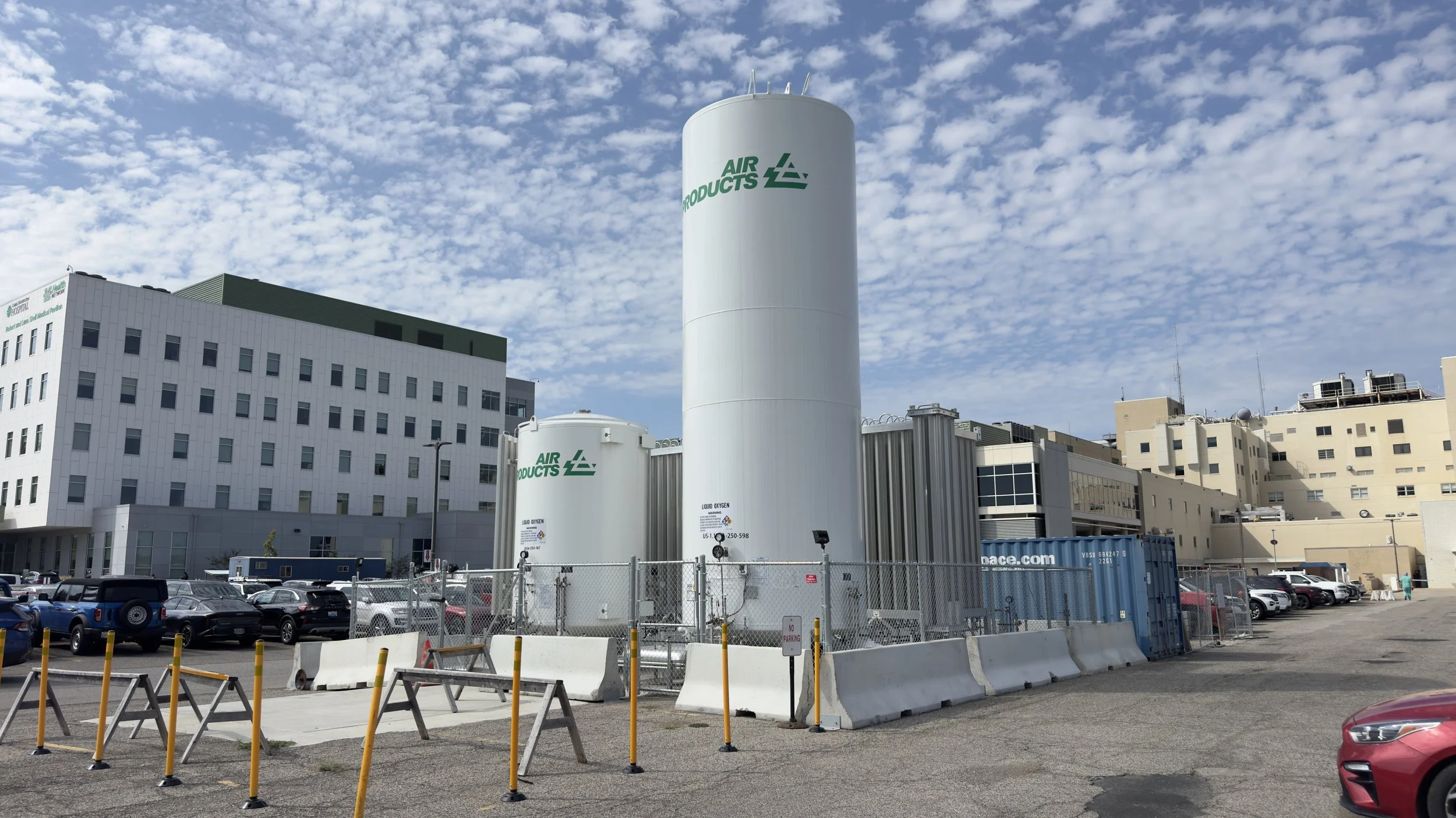 Industrial facility with large white tanks labeled 'Air Products' in a city setting, parking lot in foreground, multi-story buildings, and partly cloudy sky.