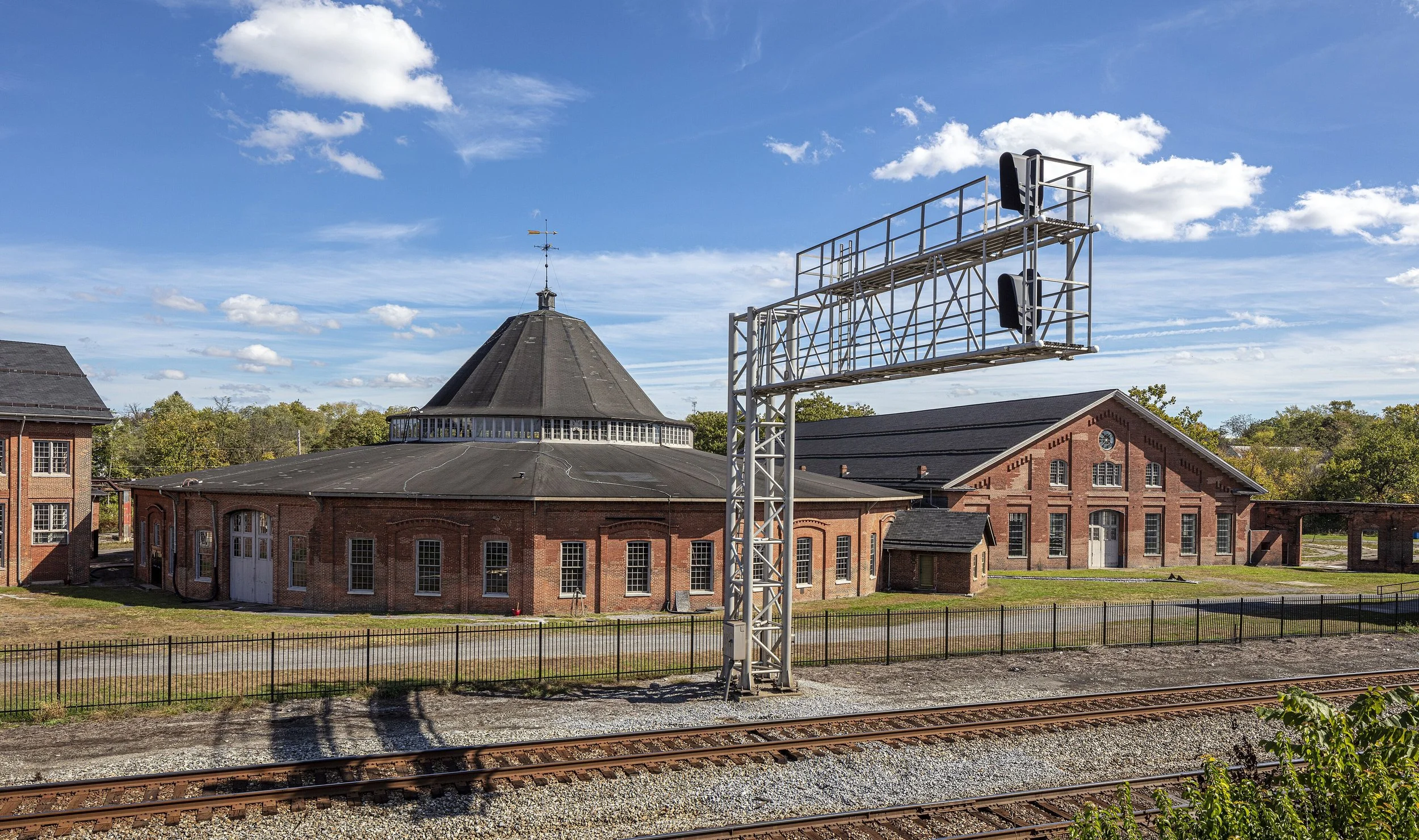 A large brick building with a conical roof and a weather vane on top. There is a metal structure with railroad signals and train tracks in the foreground, and trees under a blue sky with scattered clouds.