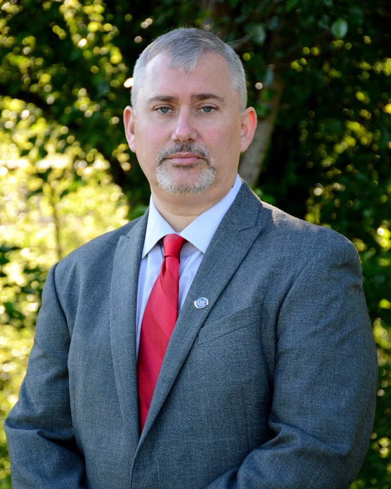 A man in a gray suit with a white shirt and red tie standing outdoors in front of green foliage.
