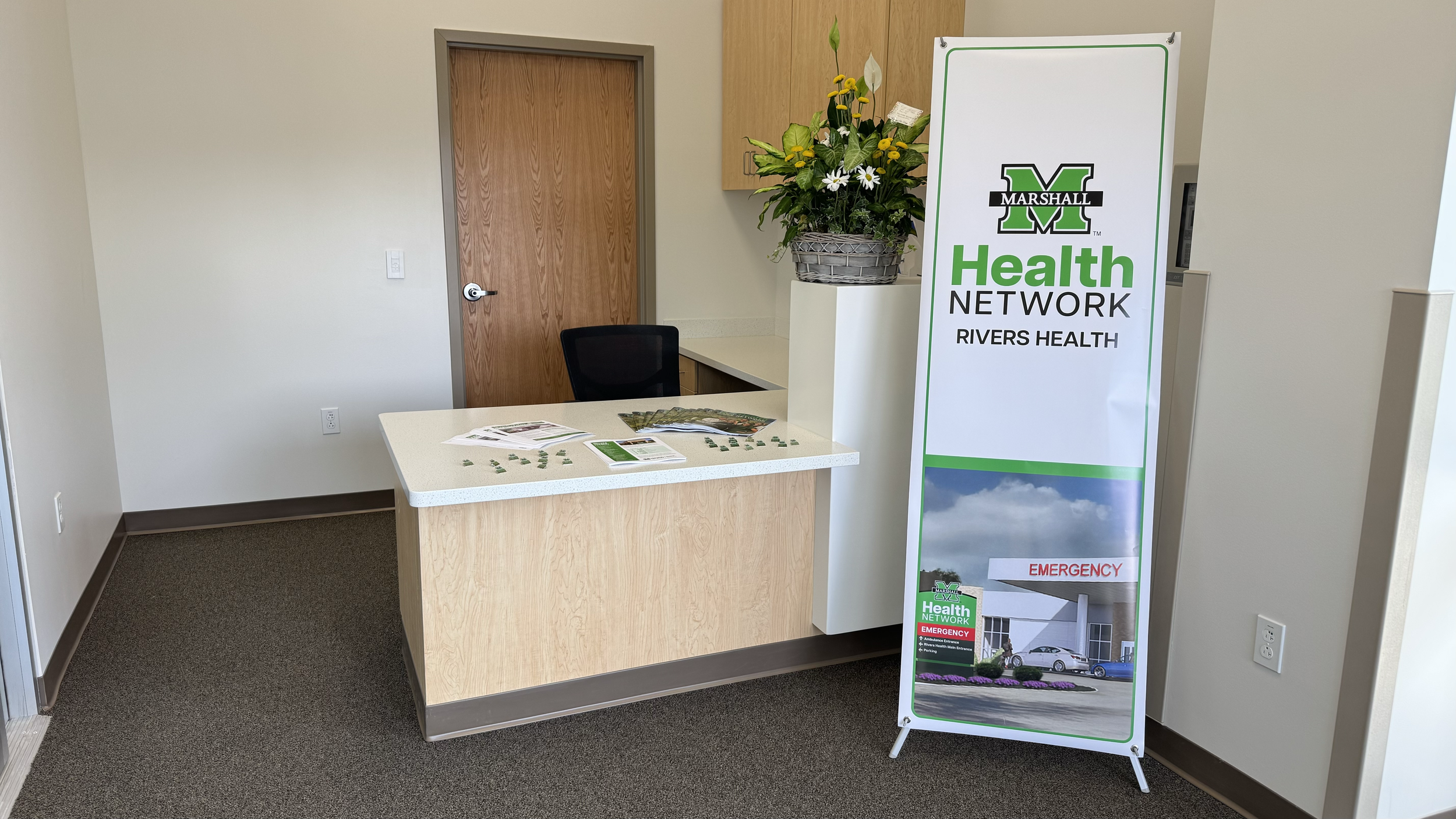 Reception area with a small table holding pamphlets, a potted plant on a counter, and a white and green health network sign for Rivers Health, Marshall Health Network with an emergency entrance photo at the bottom.