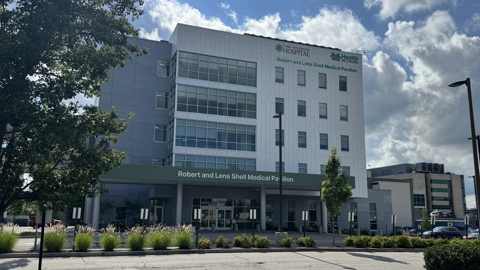 Modern multi-story hospital building with a sign reading 'Robert and Lena Shell Medical Pavilion,' surrounded by trees, shrubs, and a parking lot, under a partly cloudy sky.