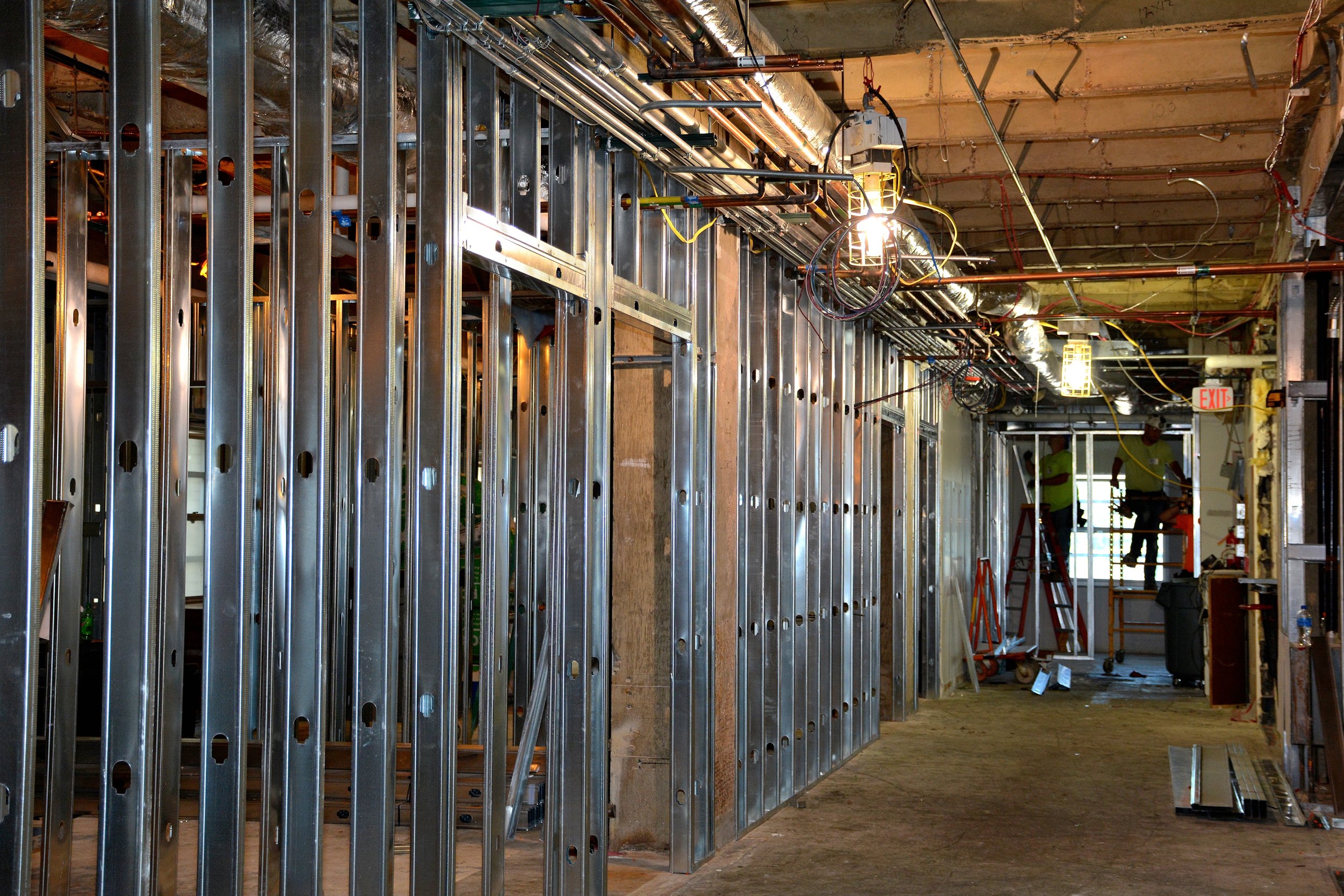 Interior construction corridor with metal stud framing, overhead ductwork, piping, electrical wiring and workers installing building systems.