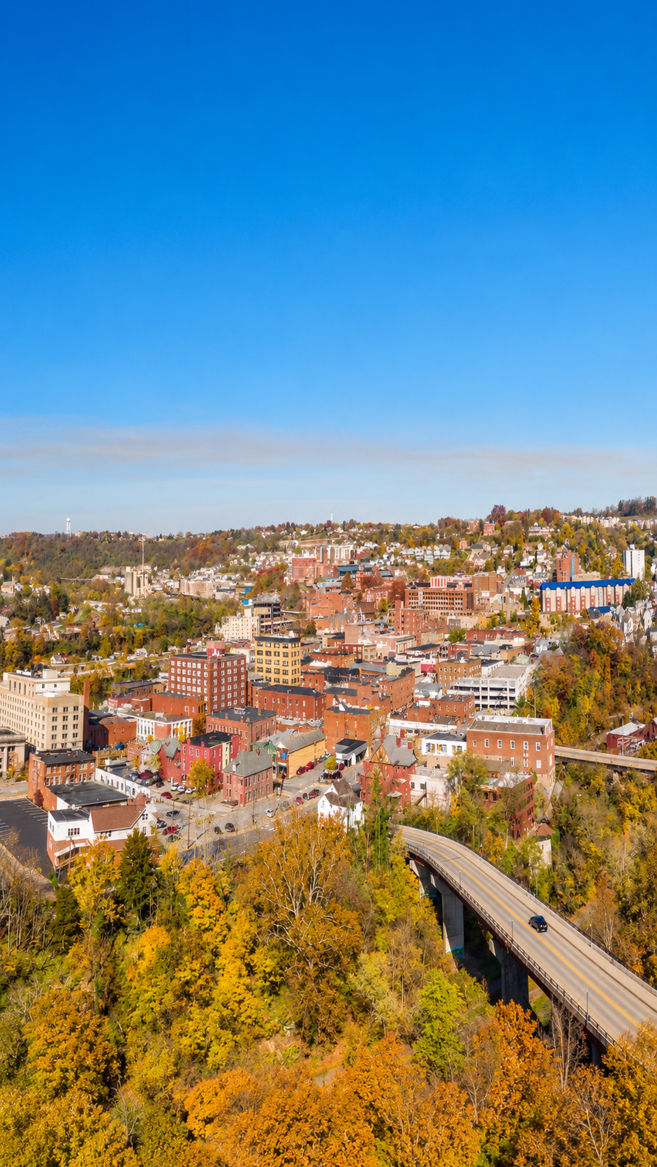 Aerial view of a city with a mix of red brick and pastel buildings, a highway with a car, and trees with fall foliage under a clear blue sky.