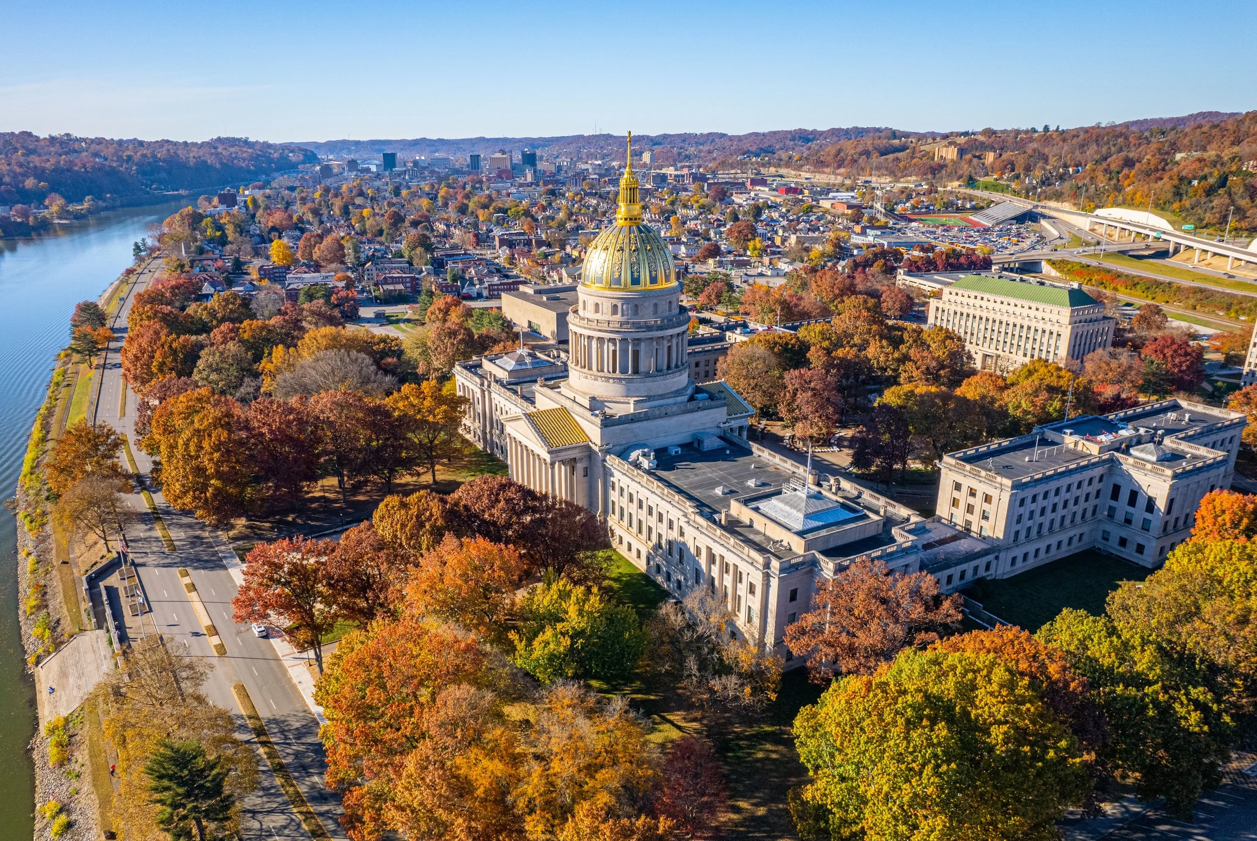 Aerial view of a city with a prominent government building with a golden dome, surrounded by colorful autumn trees, roads, and a river to the left.