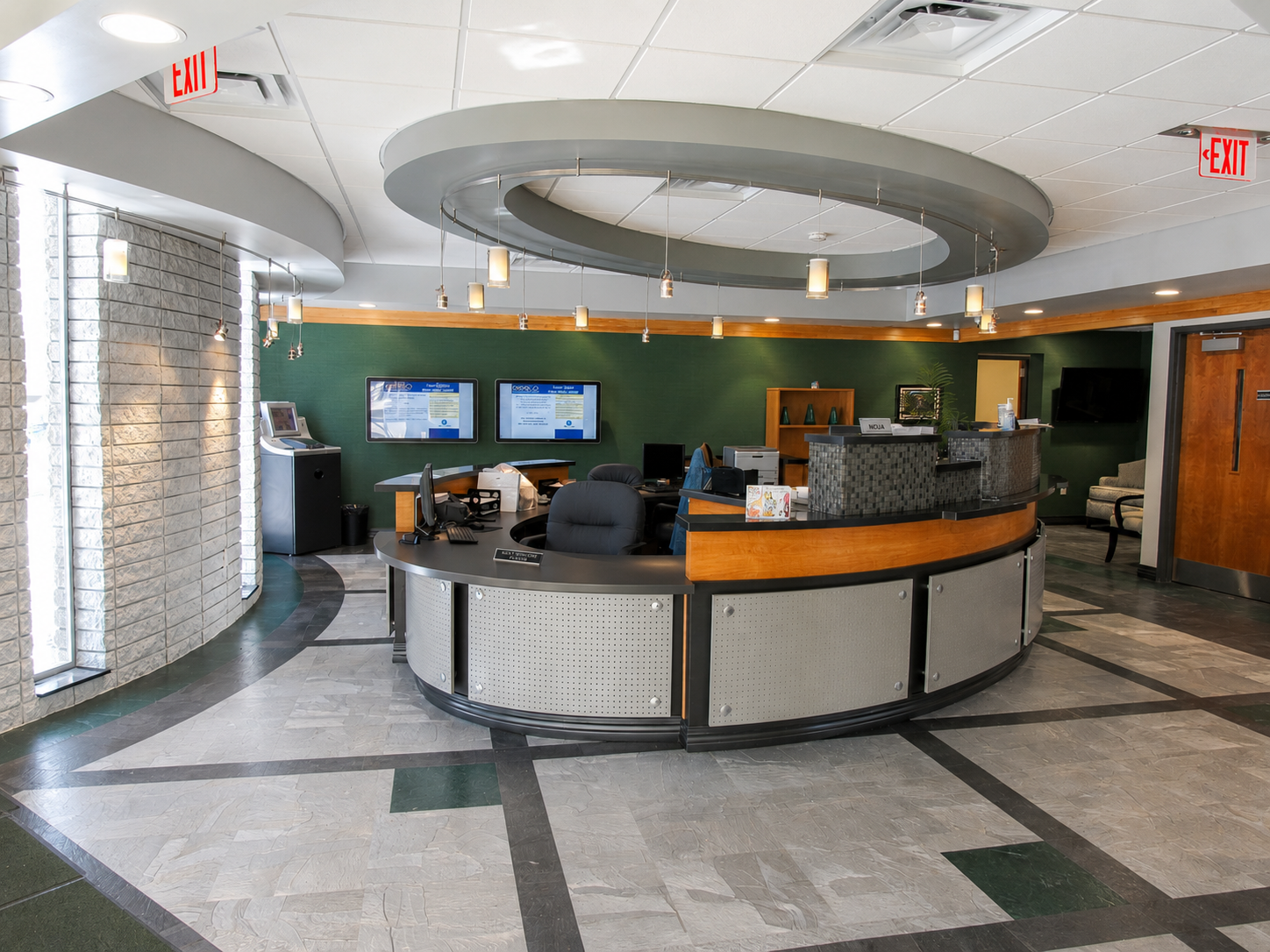 Inside a bank, featuring a curved reception desk with computer monitors, a green accent wall, wall-mounted display screens, and seating area to the right.