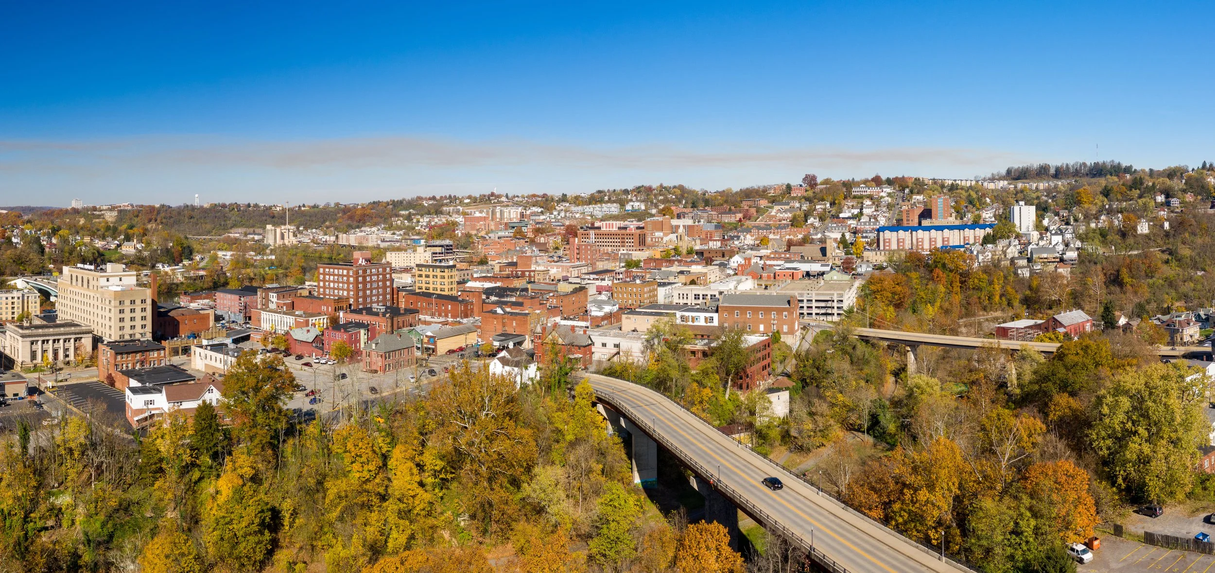 A panoramic aerial view of a cityscape with a mix of mid-rise and low-rise buildings, surrounded by trees with fall foliage. A highway with a few cars runs through the bottom right of the image under a clear blue sky.