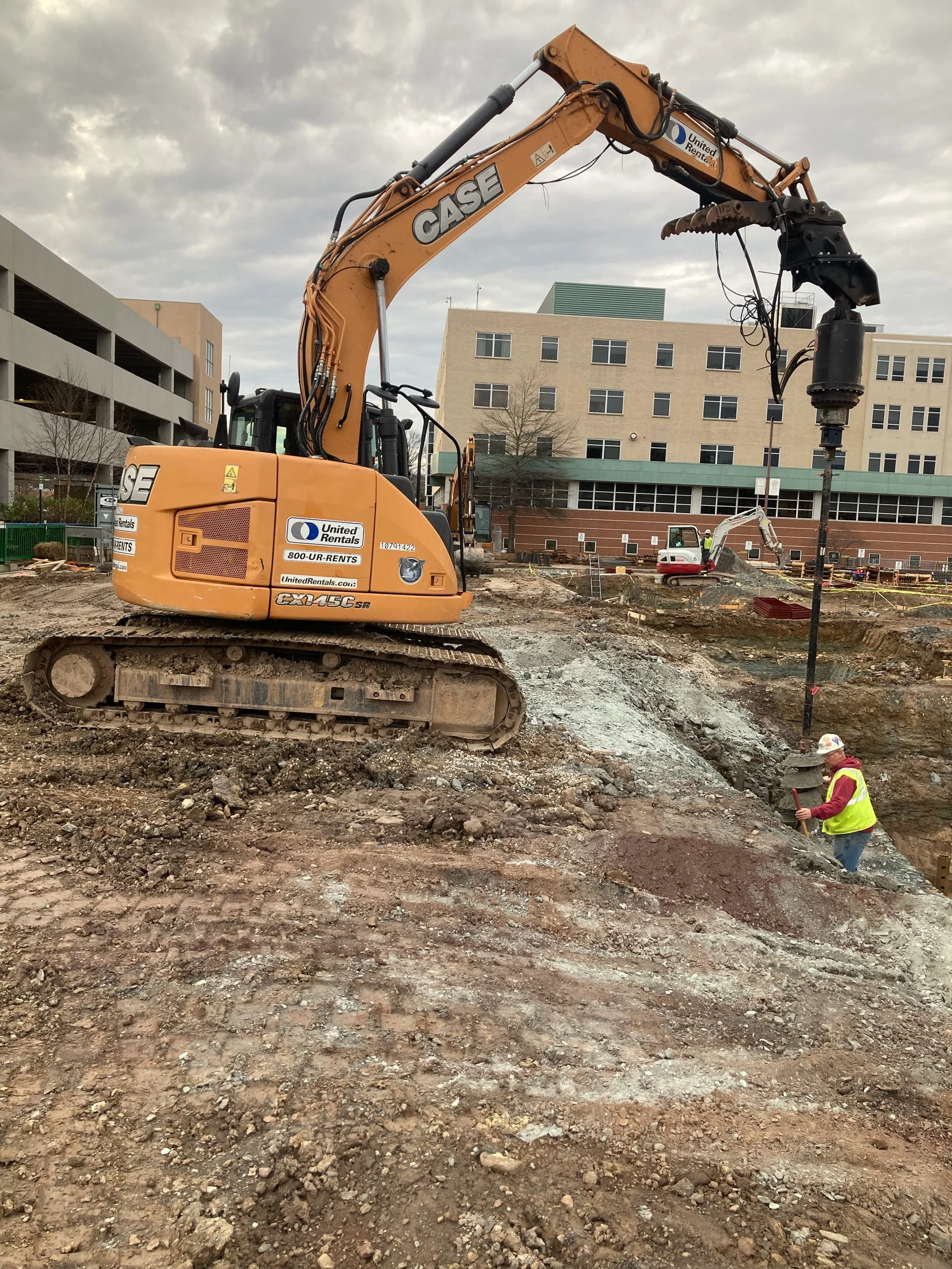 Construction site with excavator and construction worker, dirt and rocks on the ground, buildings in the background, cloudy sky overhead.