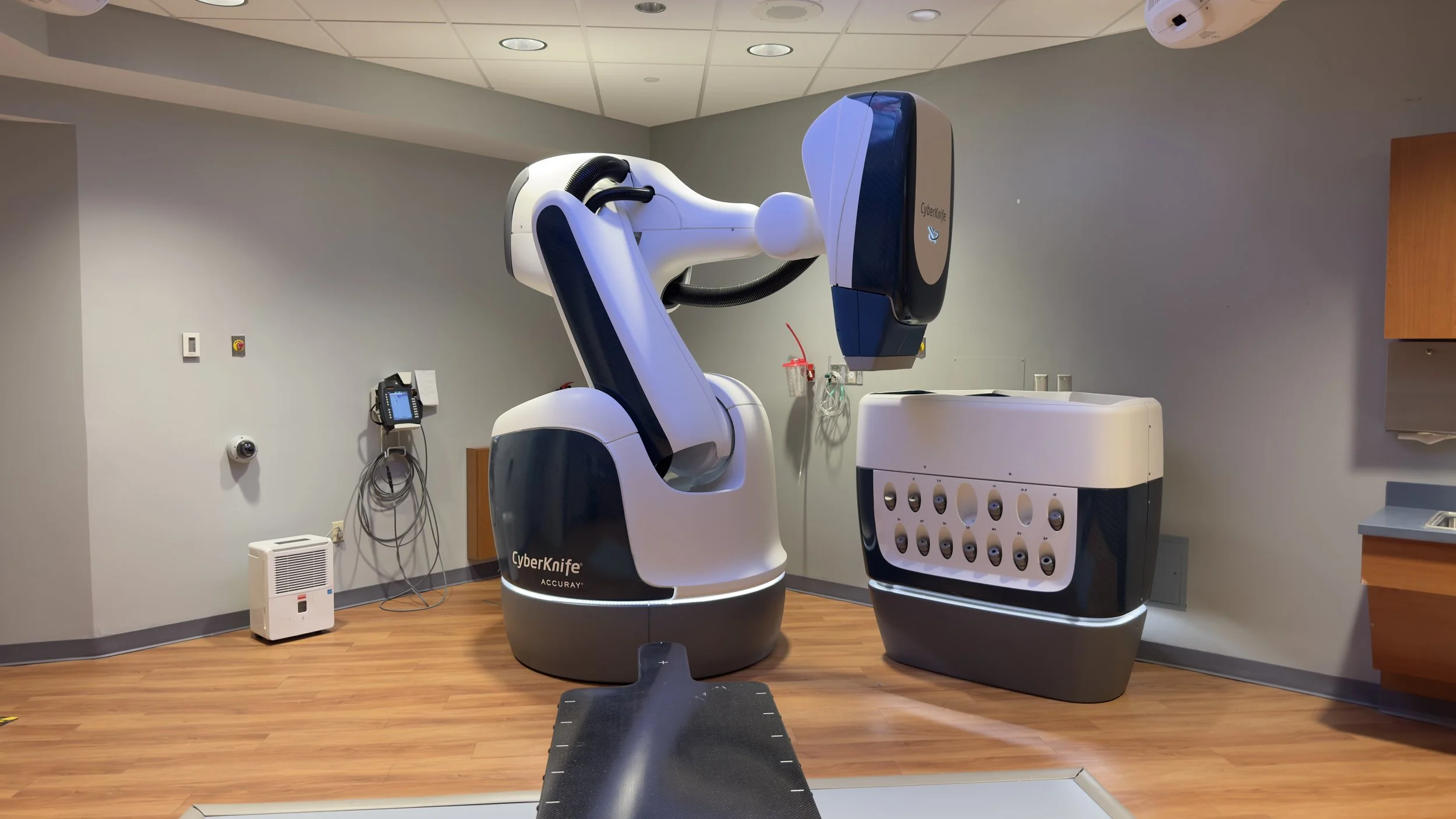 A medical room featuring a CyberKnife robotic radiotherapy system with a white and black design, a chair in front of the machine, a wall-mounted telephone, and medical equipment, all on wood flooring and grey walls.