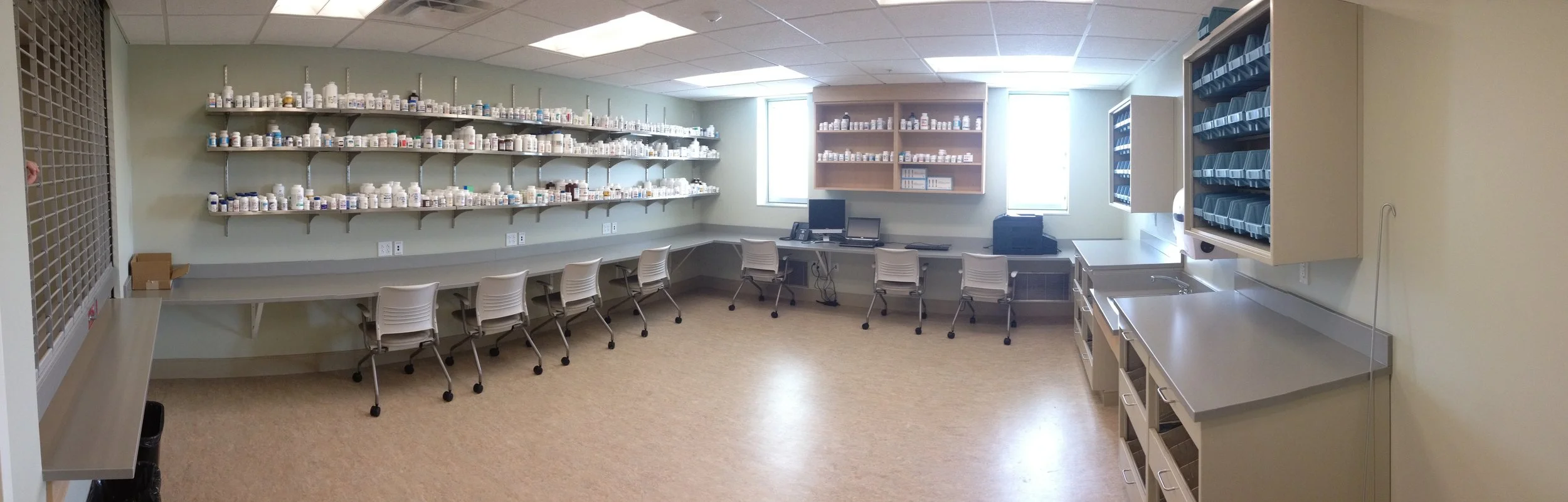A medical supplies room with shelves stocked with bottles, cabinets filled with organized trays and bins, a counter with a sink, several chairs, and a computer workstation near the window.