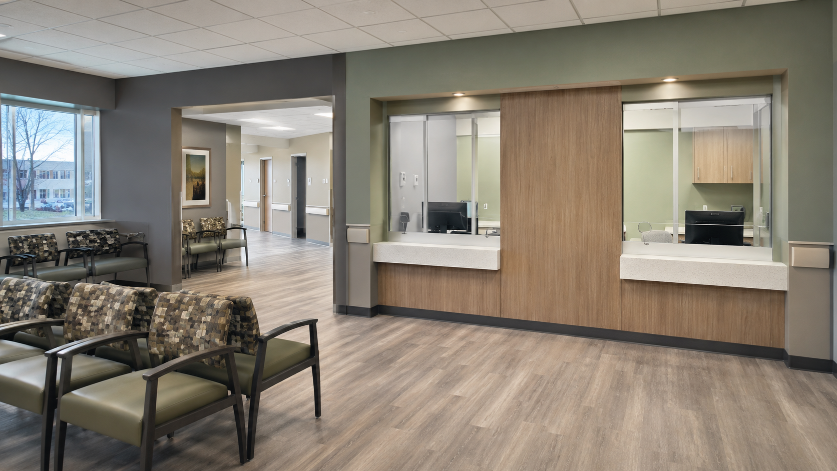 Hospital waiting area with chairs, a reception desk with two windows, and a hallway with elevators in the background.