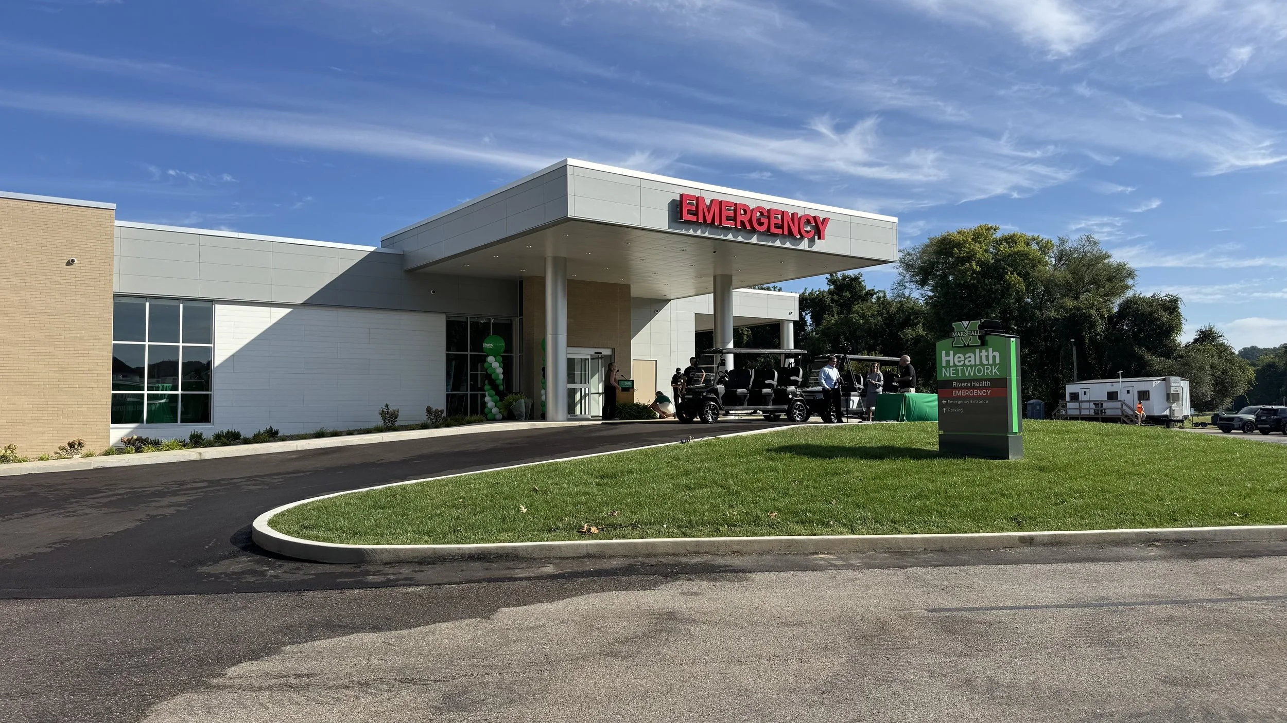 Outside of a modern emergency room building with a sign that reads 'EMERGENCY' in red letters. There are people standing near a golf cart and a large sign in the foreground indicating it is part of the Marshall Health Network. There are trees and a blue sky with some clouds in the background.