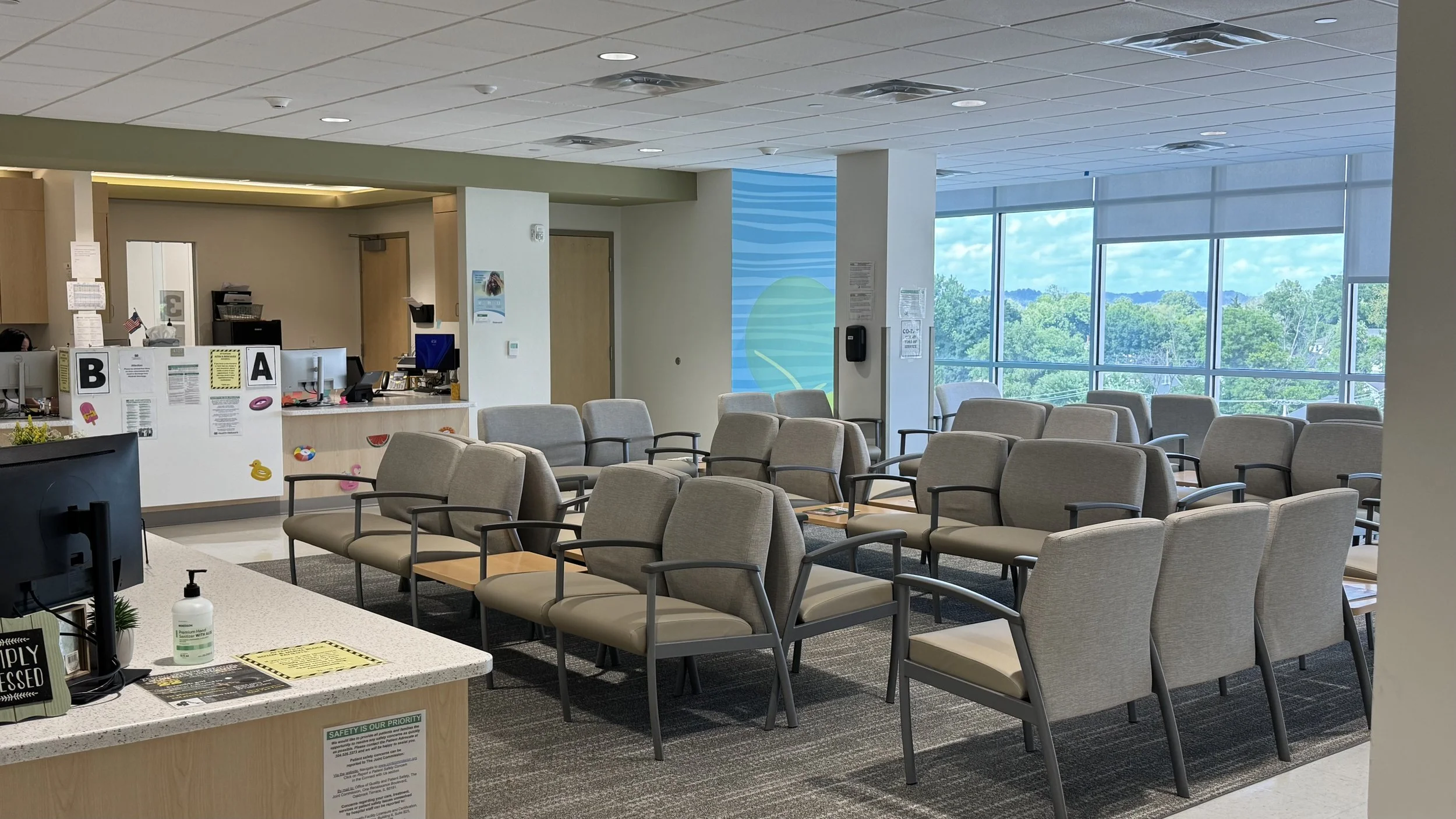Waiting area in a modern medical facility with multiple rows of beige chairs, large windows showing a green landscape, and a reception desk with signage and brochures.