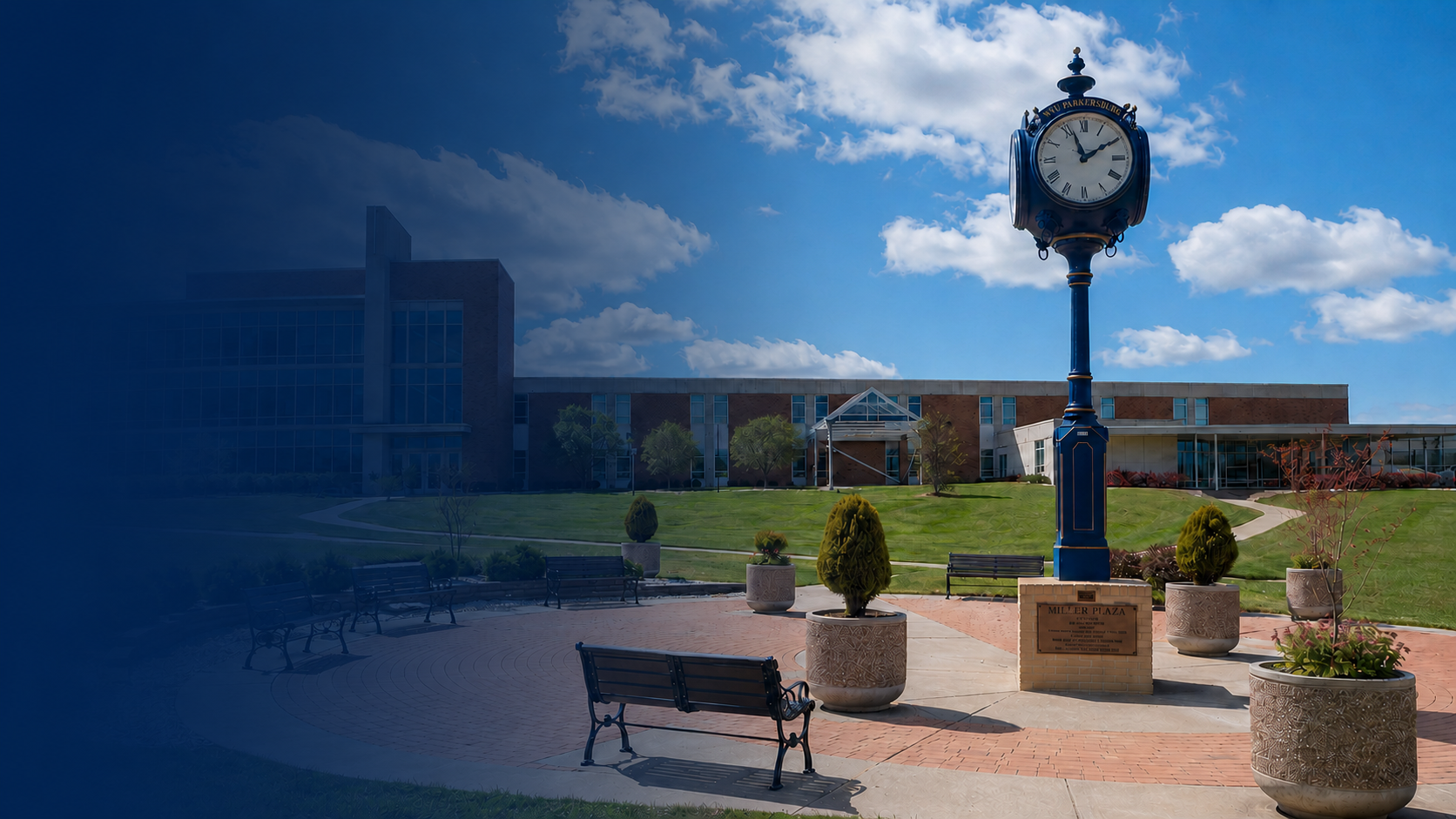 A clock tower on a brick base in a park, with benches, potted plants, and a building with glass windows in the background on a sunny day with clouds in the sky.