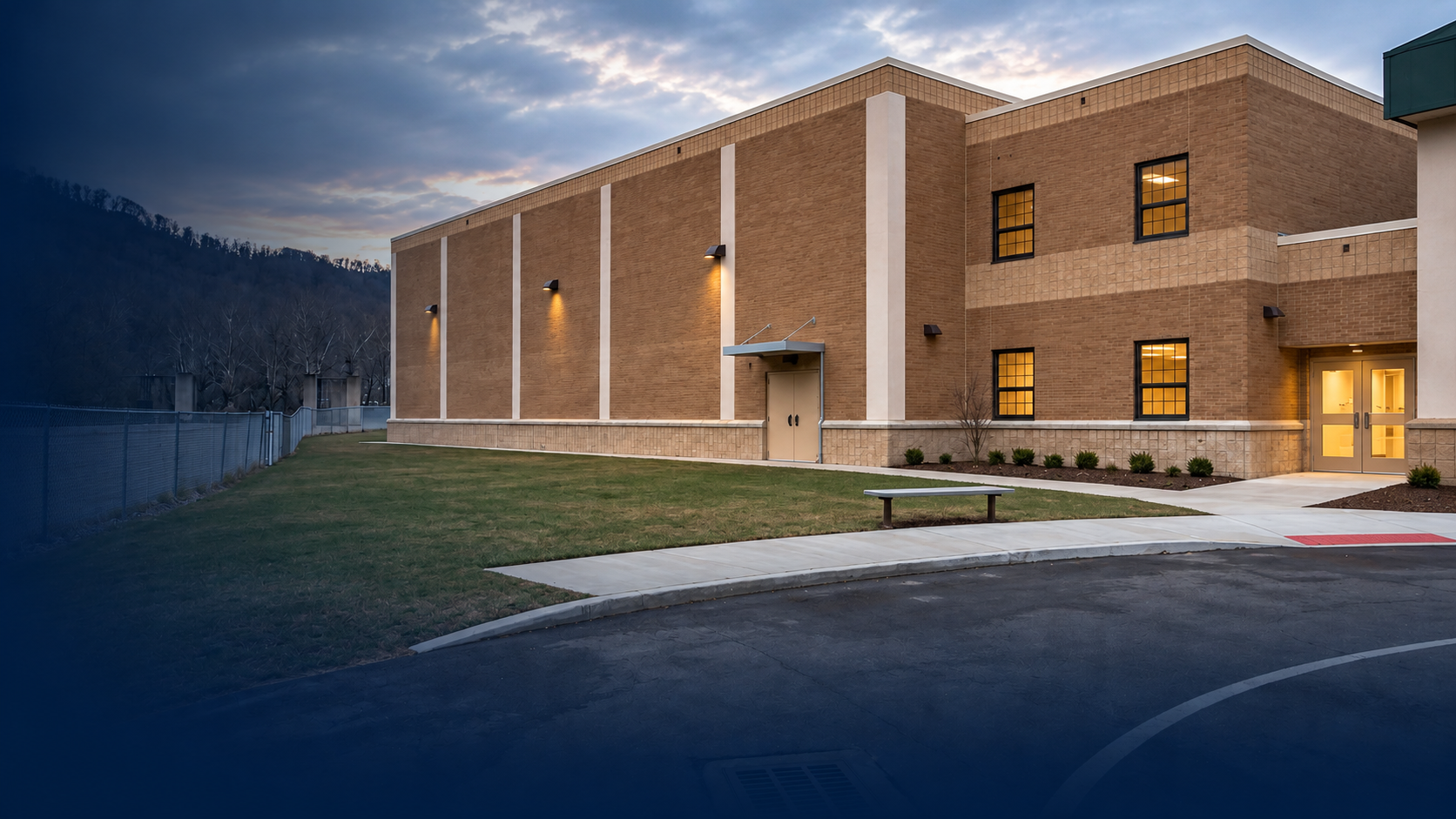 Exterior view of a modern brick building during dusk with illuminated windows, a sidewalk, grassy area, and a mountain in the background.