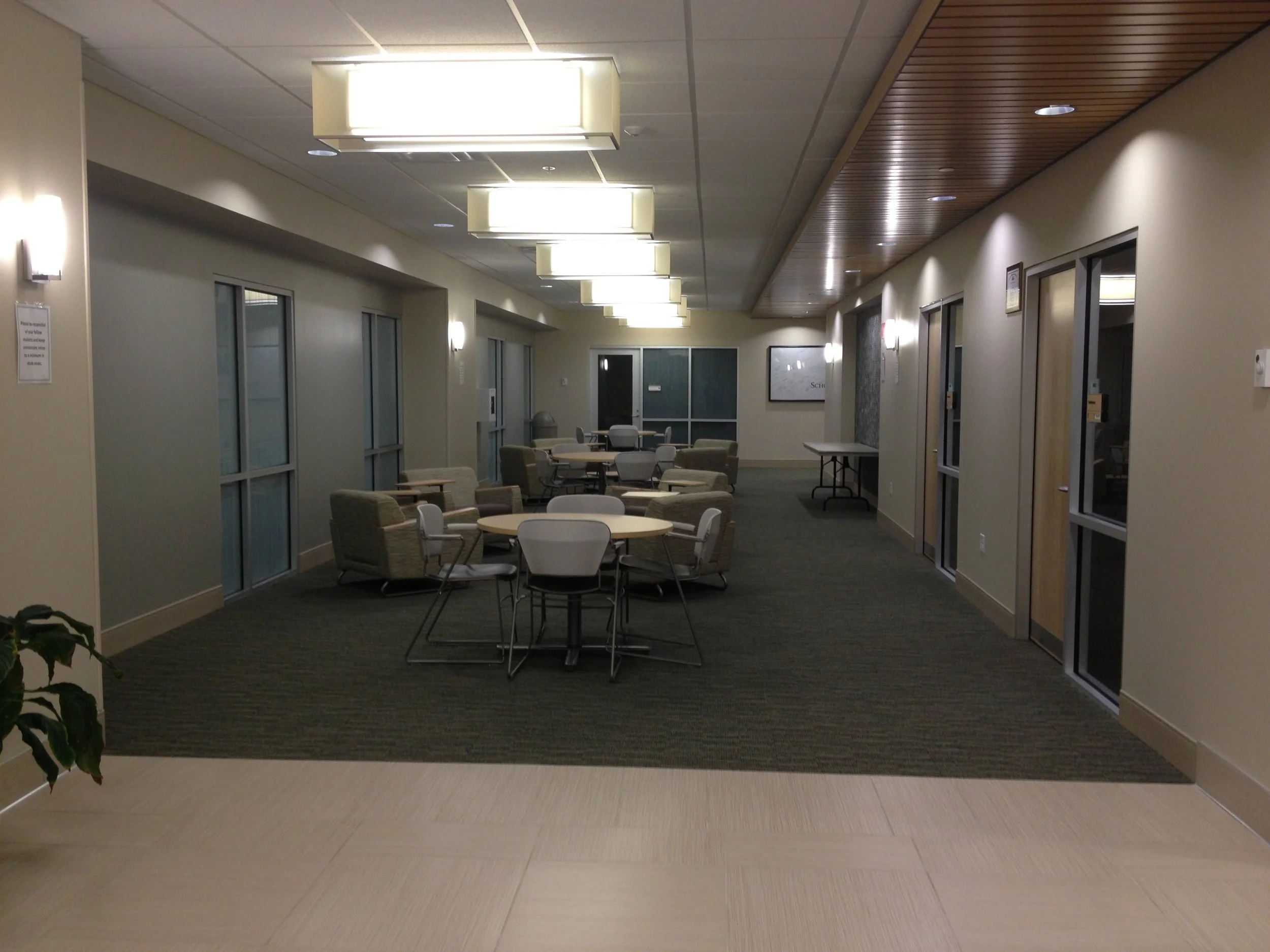 An indoor lounge area with chairs and tables, fluorescent ceiling lights, glass windows, and a wooden ceiling section.