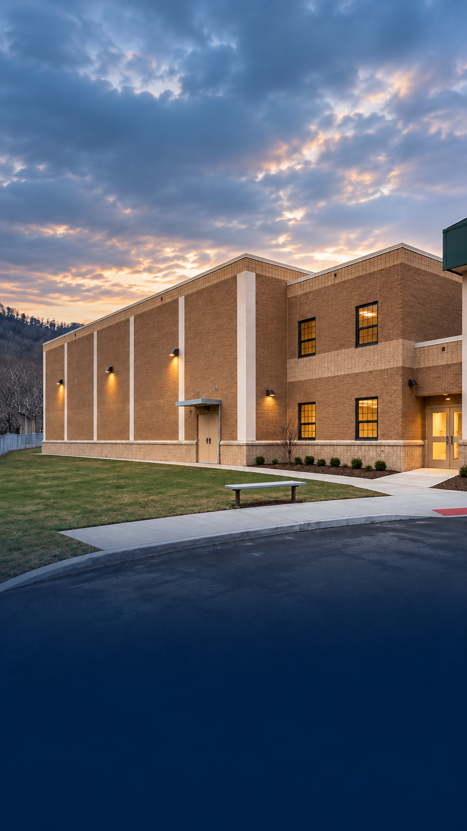 Exterior view of a Mingo County school at dusk with brick construction, illuminated windows and surrounding mountain landscape.